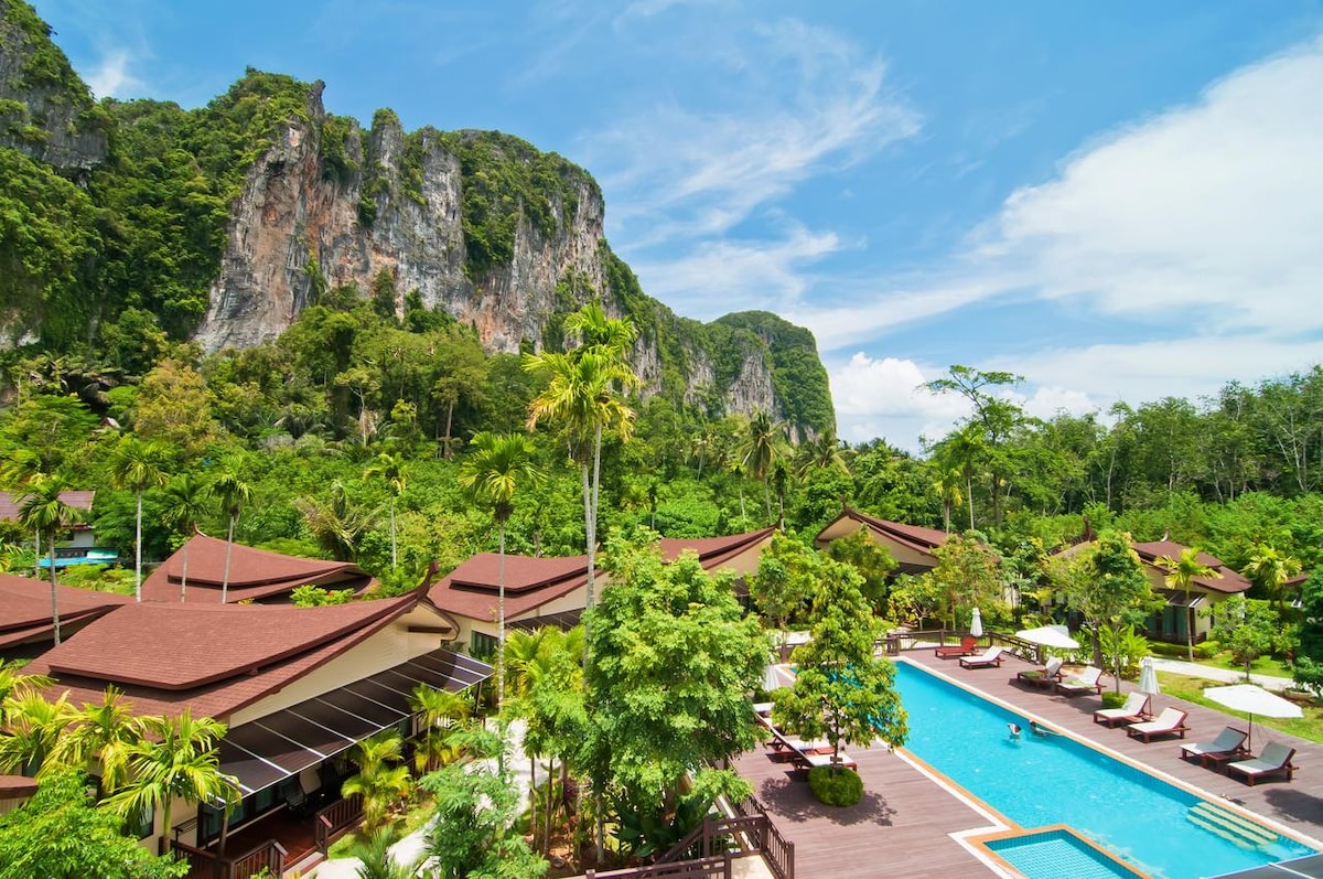 A landscaped outdoor area features a swimming pool surrounded by lush greenery and tropical plants. Sun loungers line the poolside, with views of towering cliffs in the background. The complex of bungalows is visible, nestled amid the vibrant flora.