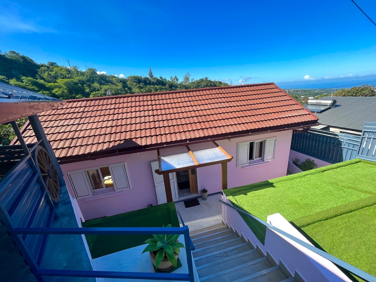 A charming pink house is seen from an elevated viewpoint, surrounded by manicured green grass. The sloped roof is adorned with terracotta tiles, and the entrance is framed by wooden beams. Scenic views of the landscape and blue sky are visible in the background.