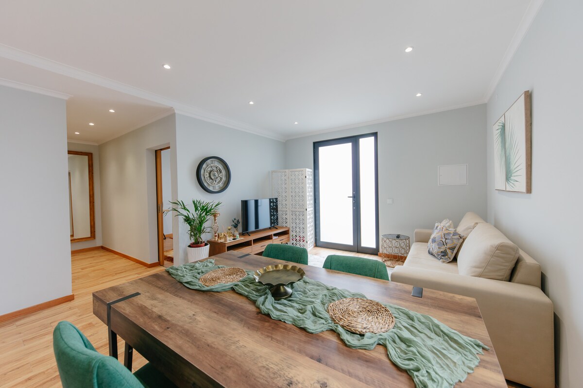 A light-filled living area features a wooden dining table surrounded by green chairs, complemented by decorative placemats. A comfortable beige sofa is arranged near a TV unit, while a potted plant adds a touch of greenery. Large glass doors lead to an outdoor space.