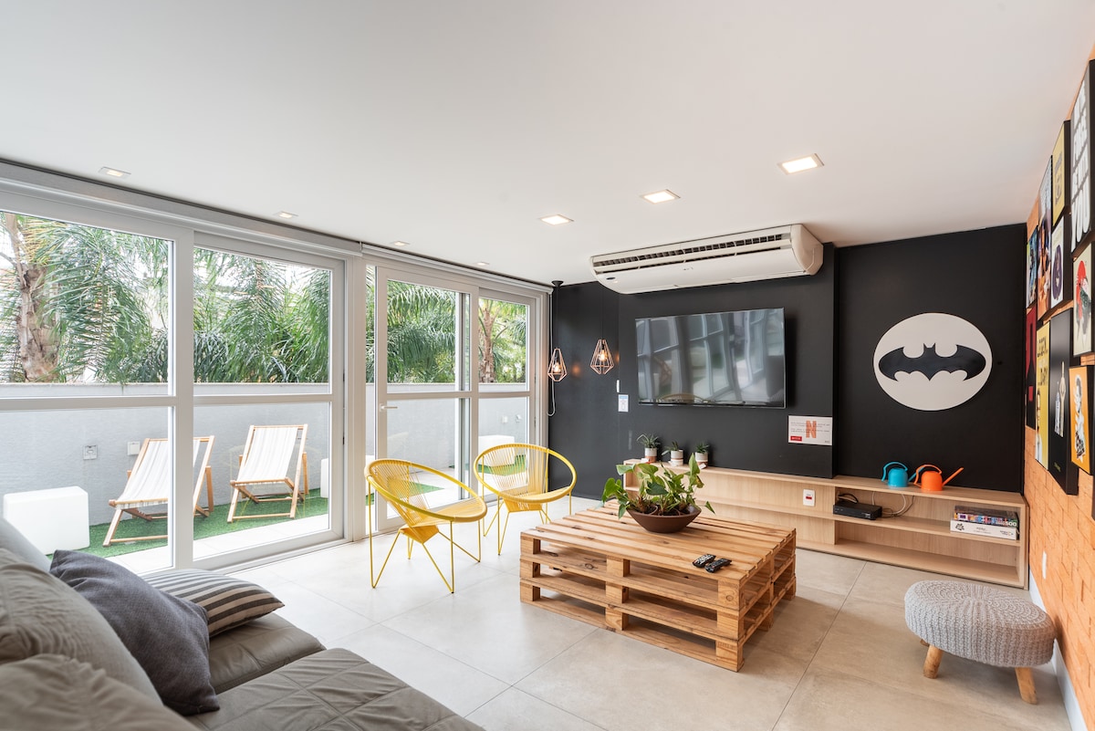 A modern living area features a wooden coffee table atop light tile flooring. Two yellow chairs provide seating, while a large television is mounted on the wall. Large windows allow natural light and view of greenery outside, enhancing the open and airy space.
