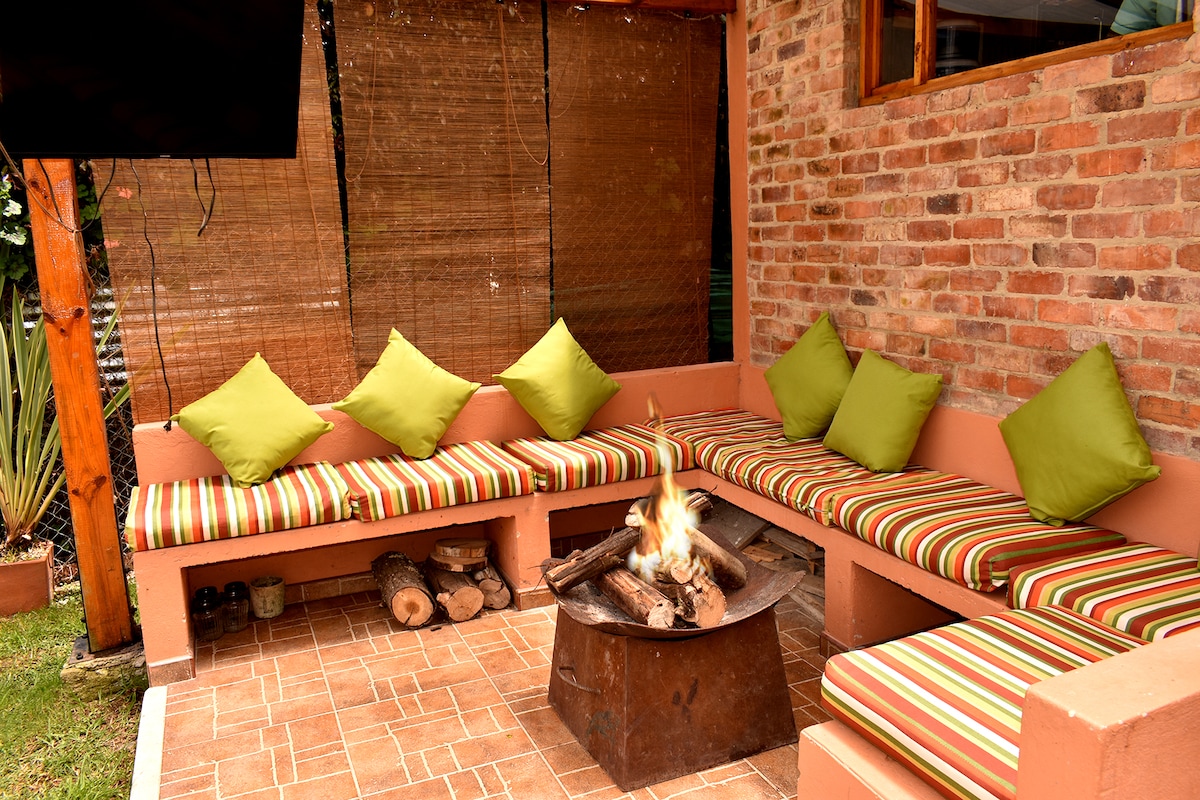 A cozy outdoor seating area is surrounded by rustic brick walls. Cushioned benches with striped fabric provide ample seating, while a fire pit is situated at the center, gathering logs for warmth. Natural light filters through the surrounding greenery, creating a relaxing atmosphere.