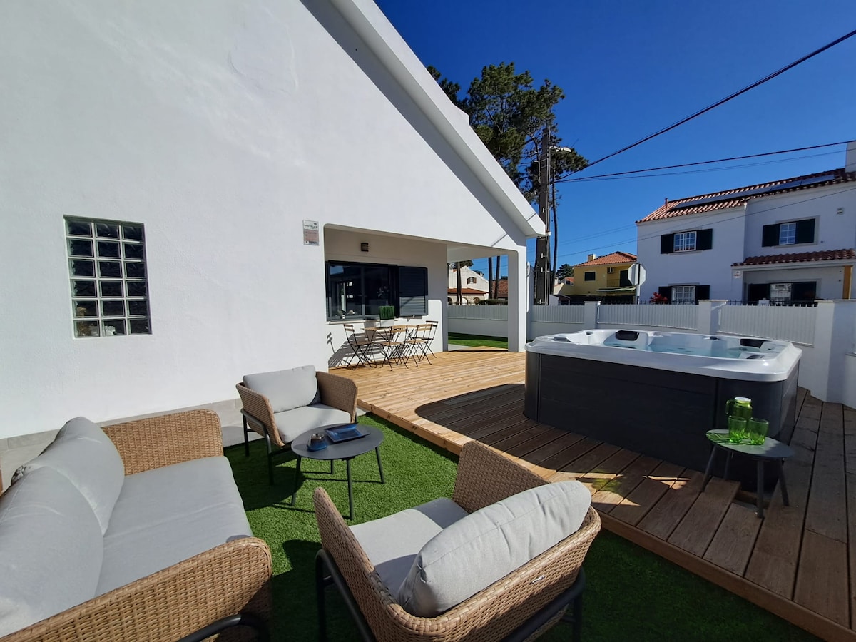 An outdoor space features a wooden deck with cozy seating arrangements. A well-appointed Jacuzzi is placed nearby, inviting relaxation. The bright blue sky overhead enhances the inviting atmosphere of the villa's exterior, complemented by green lawn areas.