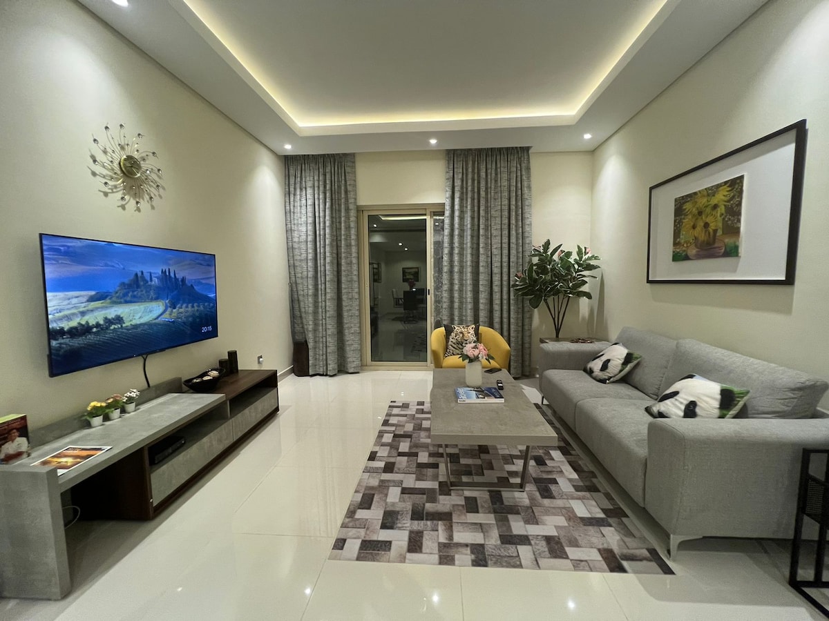 A spacious living area features a comfortable gray sofa adorned with decorative pillows, facing a large wall-mounted television. A light-colored coffee table sits on a patterned rug, complemented by lush indoor plants and large windows framed by elegant drapes.