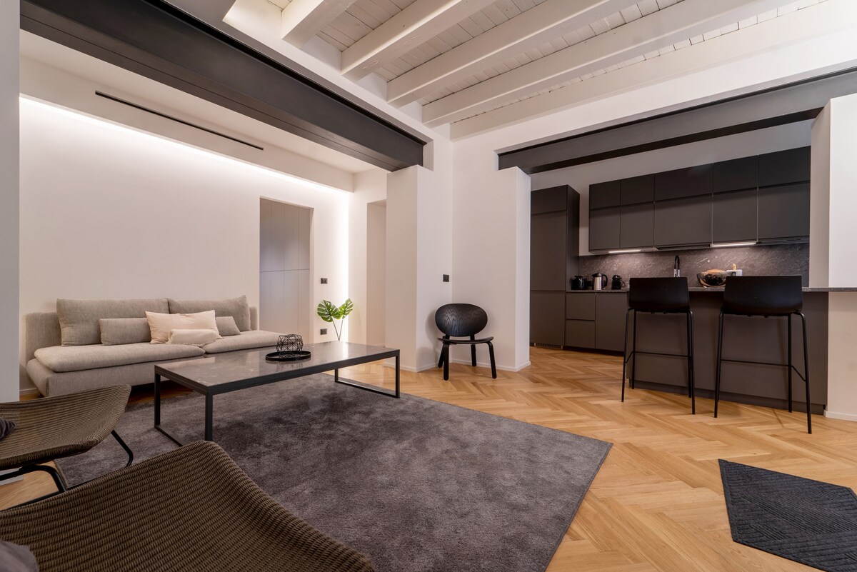A modern living area is showcased with a comfortable sofa and a low coffee table situated on a soft area rug. The kitchen, featuring sleek cabinetry and countertop, is visible in the background. Natural light illuminates the space, highlighting the refined wooden flooring.