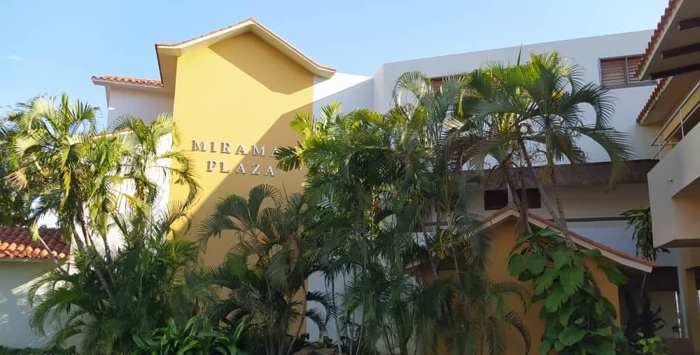 The exterior of a building is framed by lush tropical foliage, showcasing soft yellow and white walls. A sign reading 'MIRAM PLAZA' is prominently displayed on the building, while terracotta roofing adds a warm touch to the architectural style.