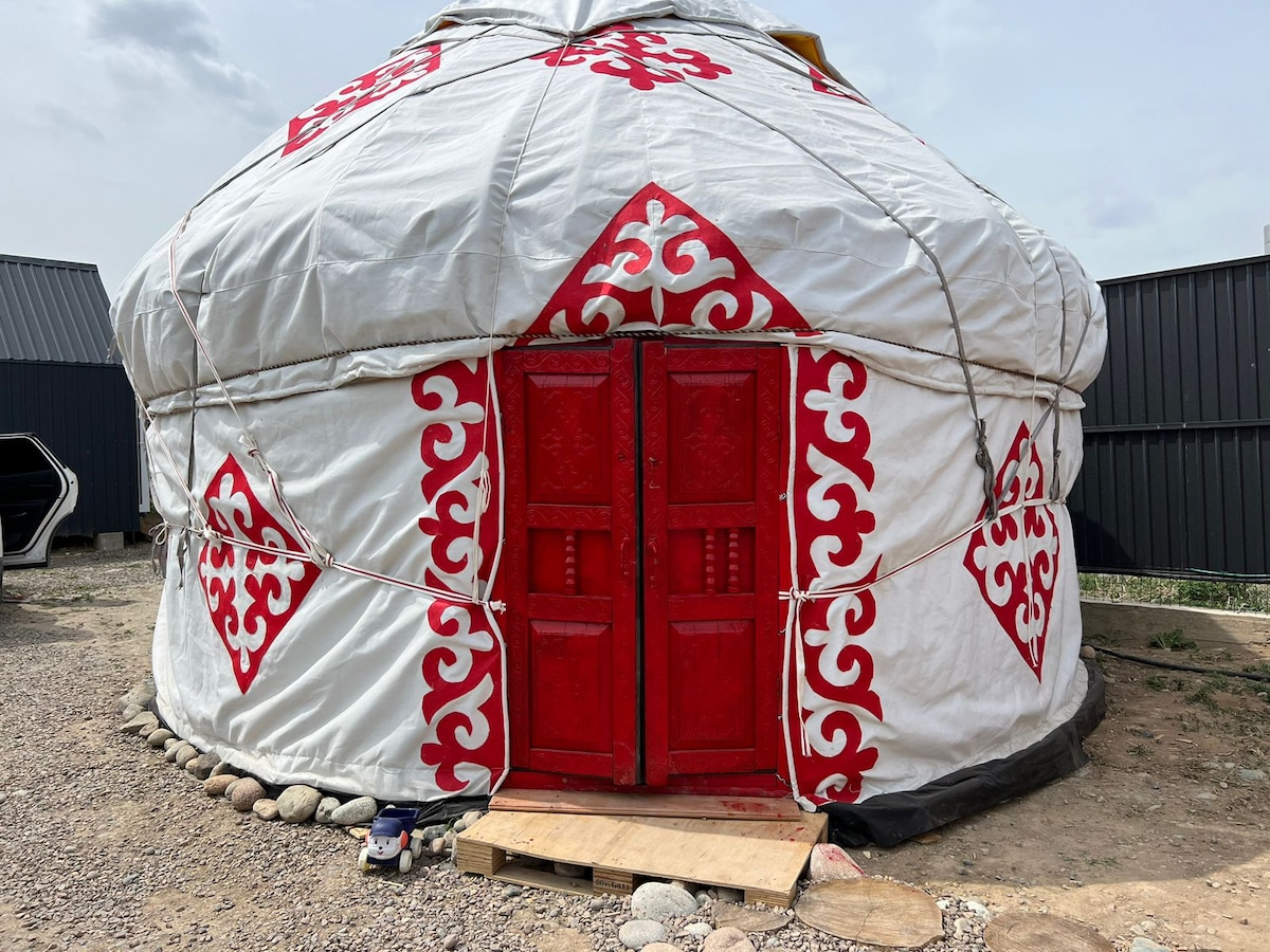 A traditional yurt is featured, showcasing its circular shape and distinctive white exterior adorned with red ornamental patterns. The entrance is marked by double wooden doors, painted in a vibrant red. A compact wooden platform is positioned at the doorway.