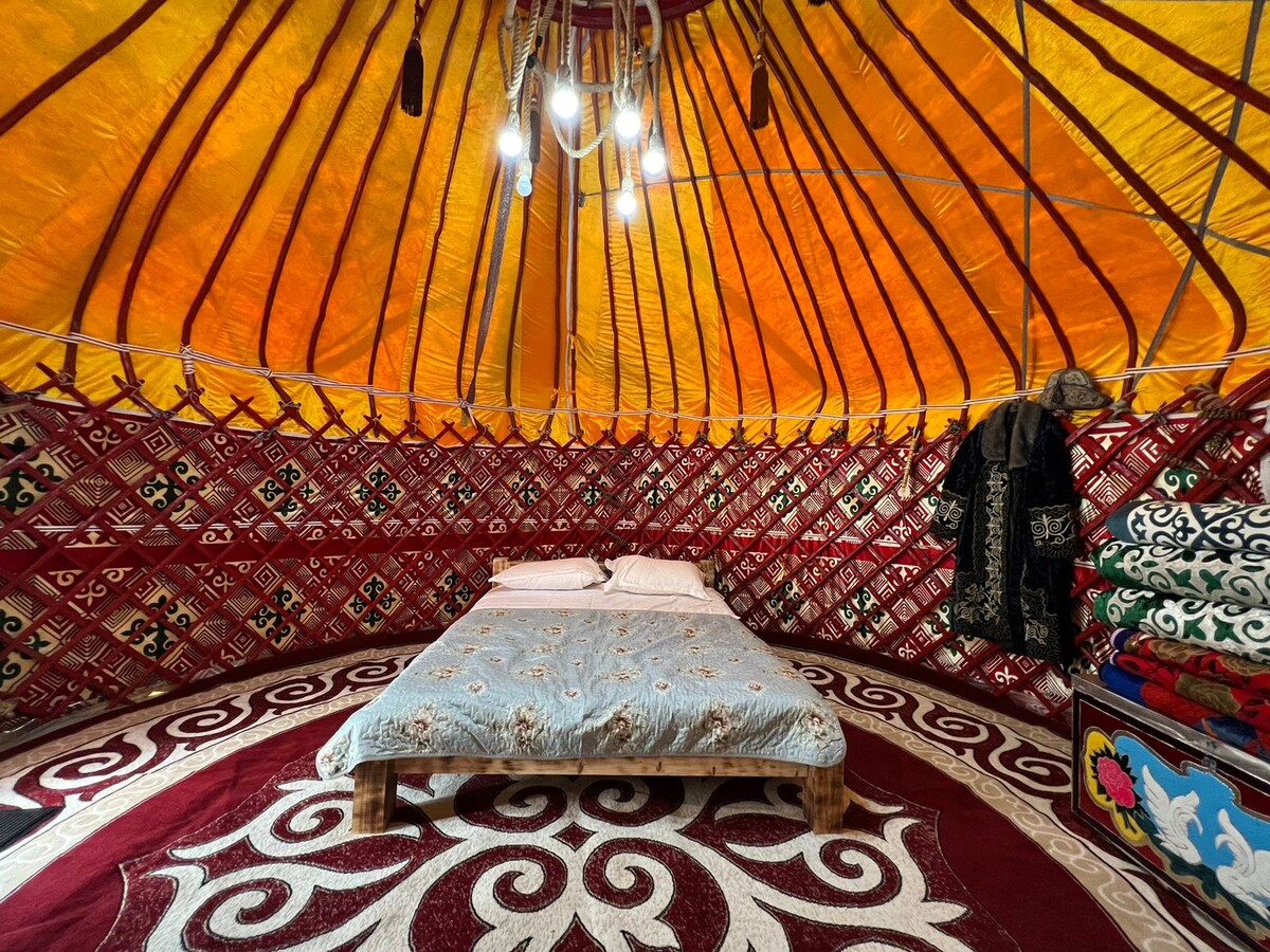 The interior of the yurt features a spacious wooden bed with layered bedding. Colorful wall coverings made of traditional patterns surround the space, complemented by a patterned carpet on the floor. Bright lights hang from the ceiling, adding warmth to the environment.