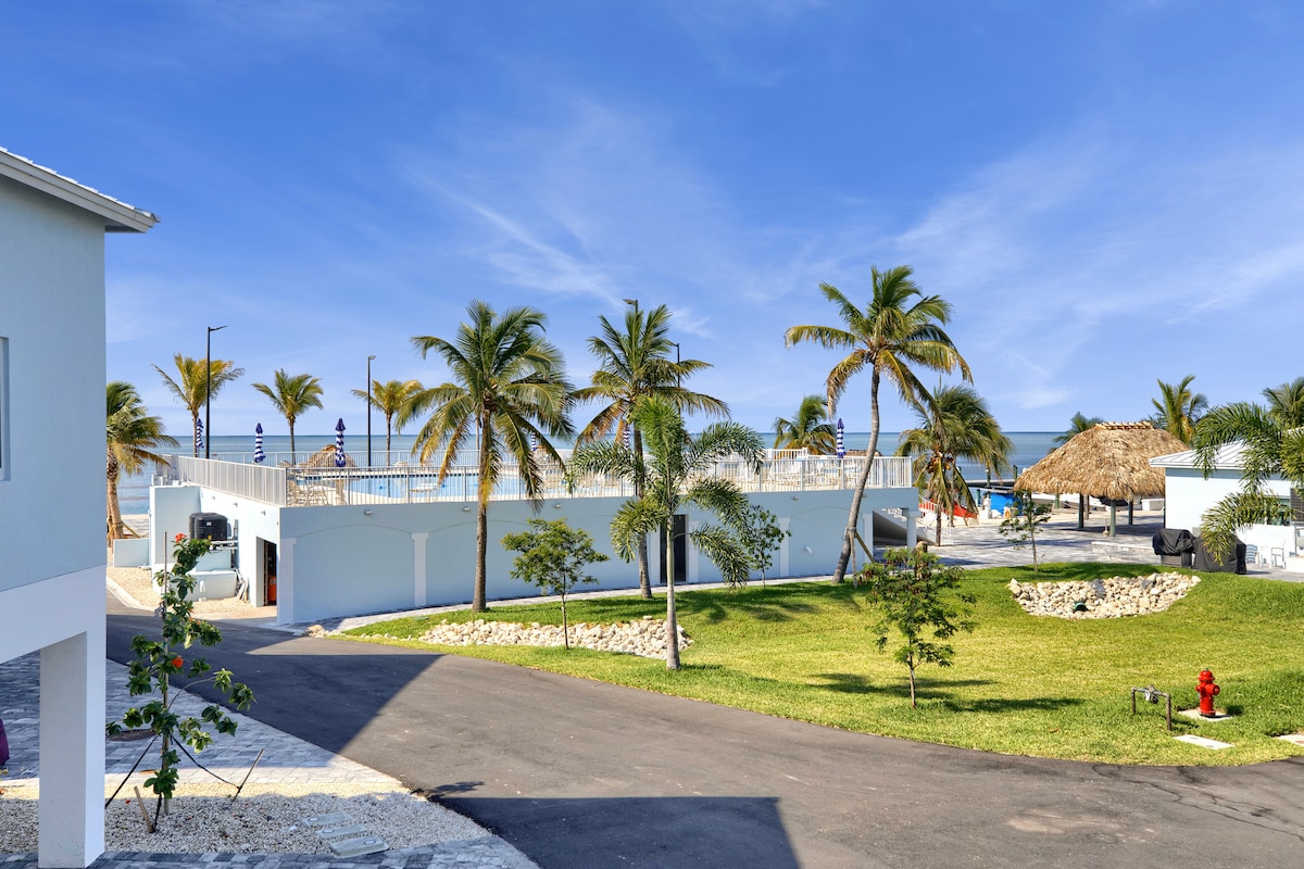 A clear view of the property’s exterior, featuring neatly maintained green lawns and tropical palm trees. The elevated swimming pool is visible alongside shaded Tiki huts, with the ocean in the background. The pathway leads towards the building with accessible parking areas.