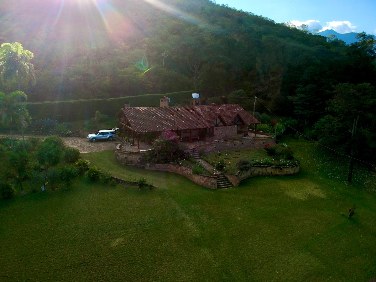 A spacious cabin is nestled within a lush green landscape, surrounded by trees and mountain views. The property features well-maintained gardens with vibrant flowers, while a vehicle is parked nearby. Sunlight filters softly through the trees, enhancing the tranquil outdoor setting.