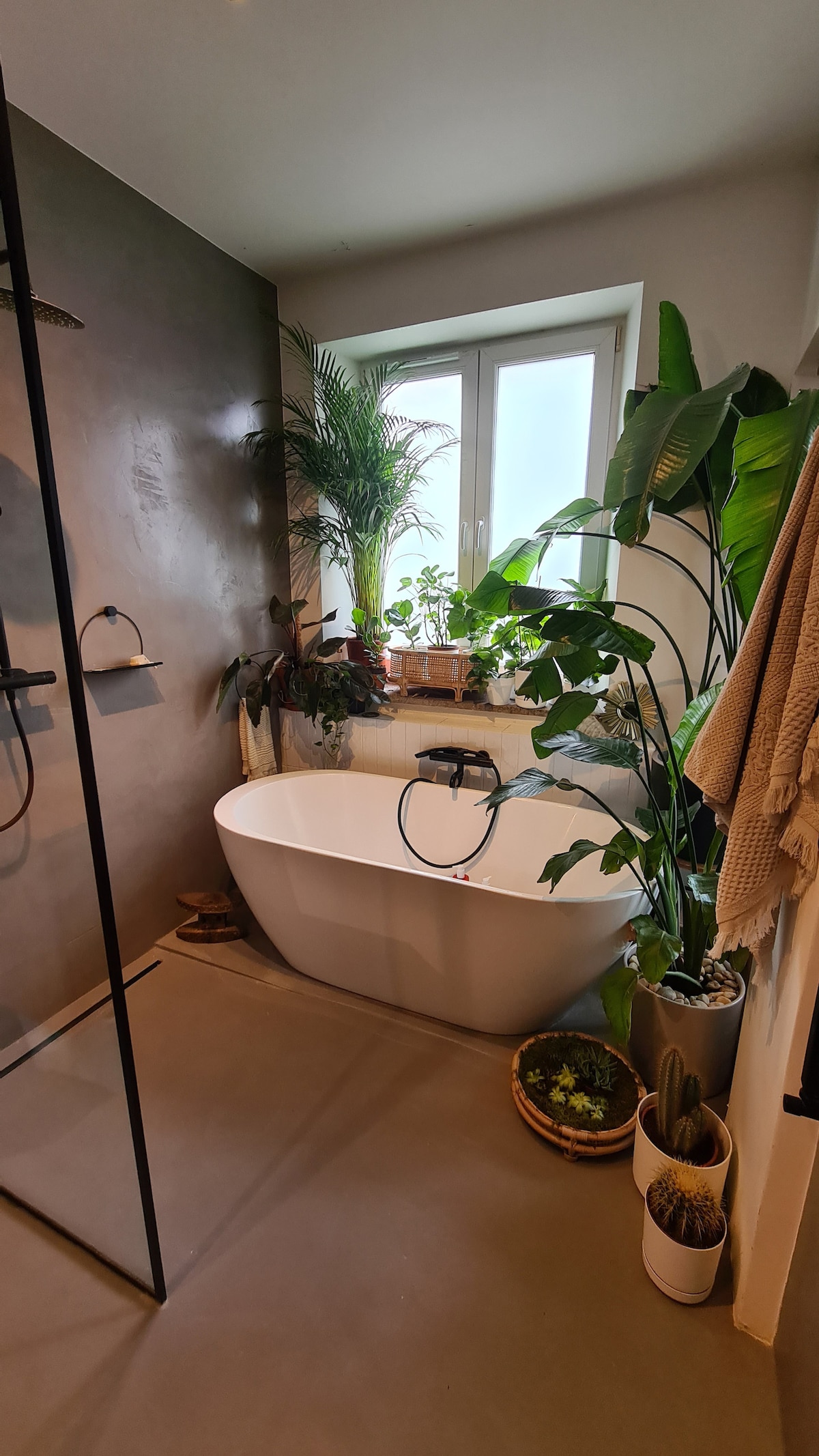 A modern bathroom features a freestanding bathtub positioned next to a window adorned with greenery. Various plants are positioned on the counter and floor, contributing to a calm environment. Light filters in through the window, complementing the neutral tones of the walls and flooring.