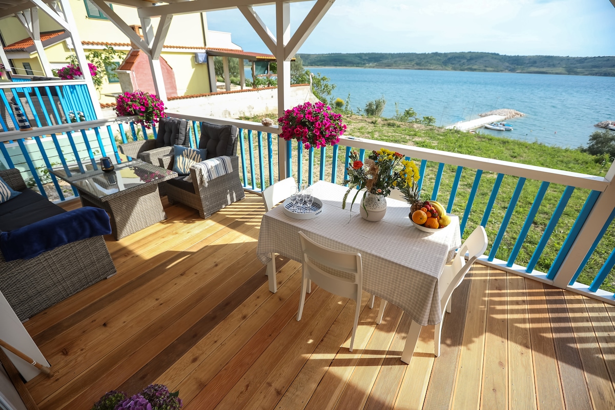 A spacious terrace features a dining table set with a tablecloth and fresh fruit, surrounded by comfortable seating options. Vibrant flowers in pots add color to the area, while the serene view of the sea and lush greenery creates a peaceful atmosphere.