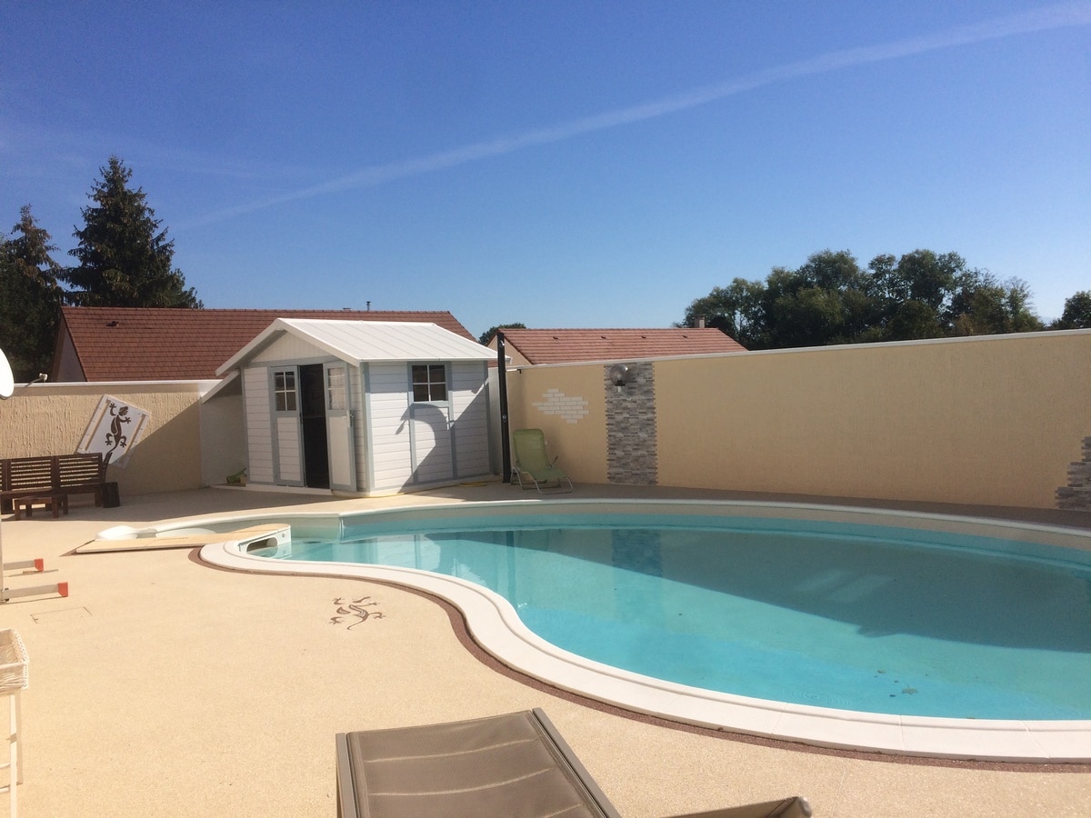 A private swimming pool is surrounded by a sun deck with loungers, offering a relaxing space. A shed is positioned near the pool, with a light-colored exterior and decorative stone accents. The area is enclosed by a smooth, high wall, providing privacy.