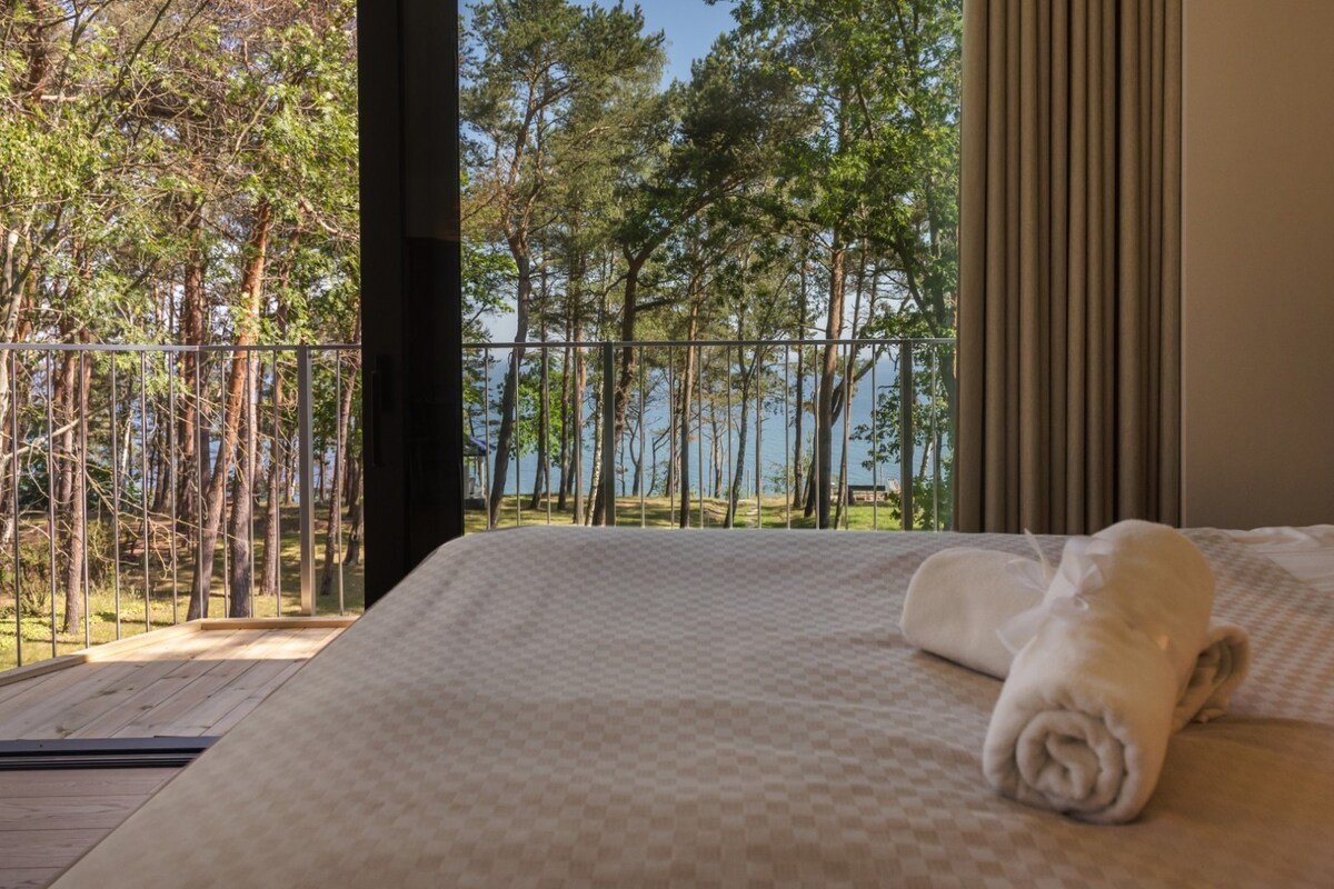 A room features a spacious bed adorned with a rolled towel, positioned next to large glass doors. The view reveals a lush forest of trees leading to a serene body of water, providing an expansive sense of nature from the indoor space.