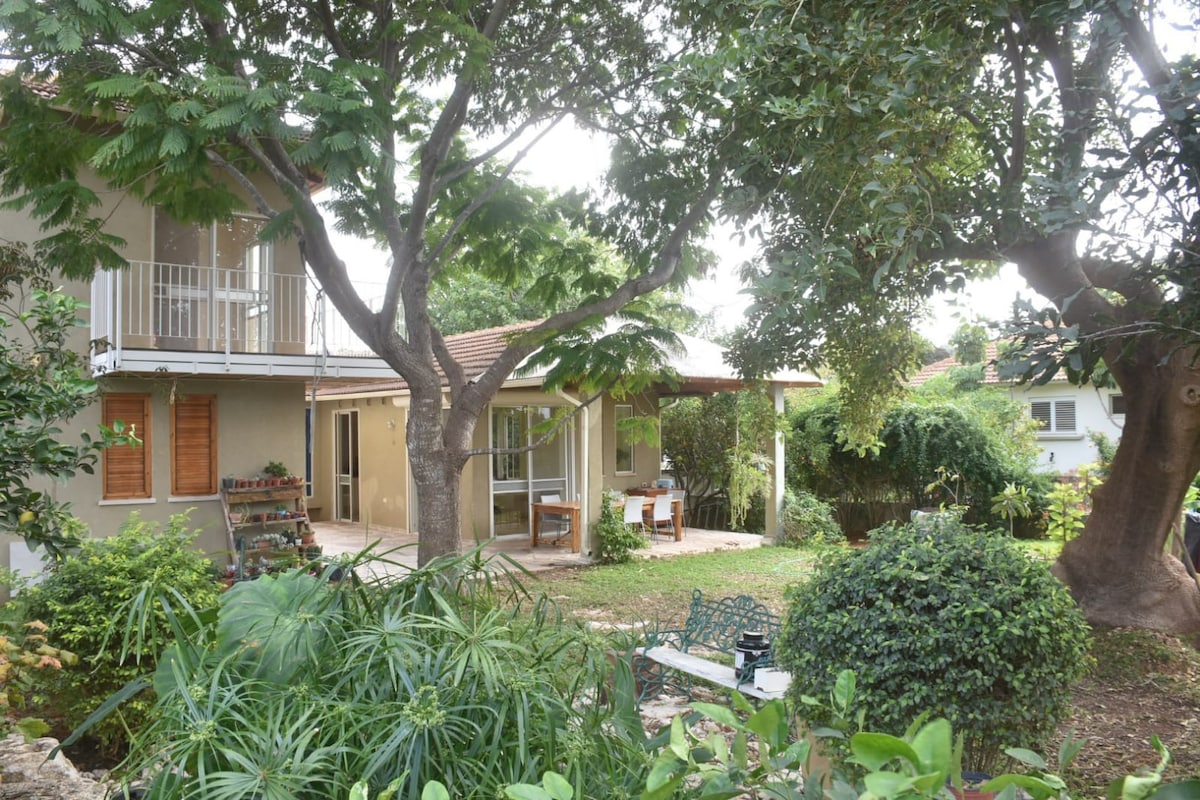 A serene outdoor area surrounds the house, featuring lush greenery and mature trees. A spacious patio is visible, with outdoor seating and tables, inviting gatherings. The design offers an atmosphere of tranquility, with various plants enhancing the natural setting.