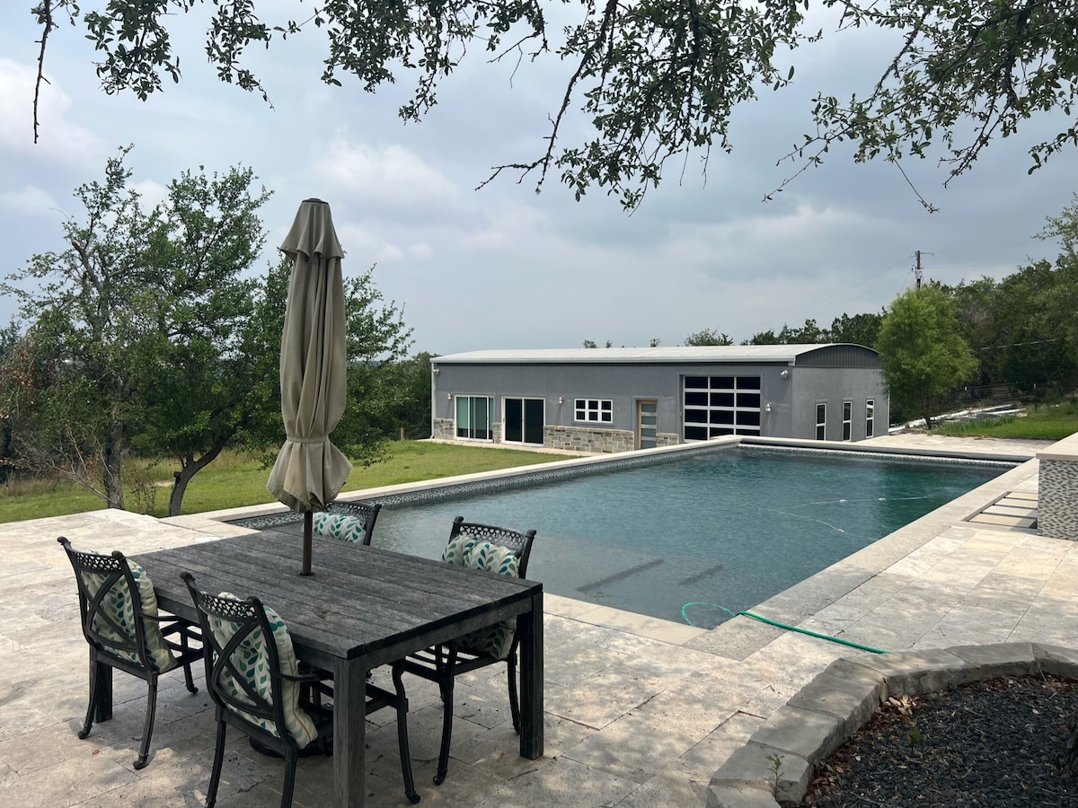 An outdoor space showcases a large pool surrounded by a stone patio. A dining table with chairs is positioned nearby, complemented by a shaded umbrella. In the background, a modern guesthouse is visible, with large windows reflecting the surrounding greenery and landscape.
