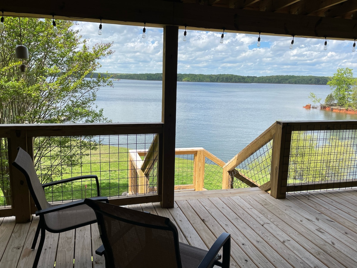 A covered deck offers a view of the calm water, framed by trees. Two chairs are positioned on the wooden deck, with steps leading down to the grassy shoreline. Soft lighting hangs along the deck, providing a welcoming ambiance.
