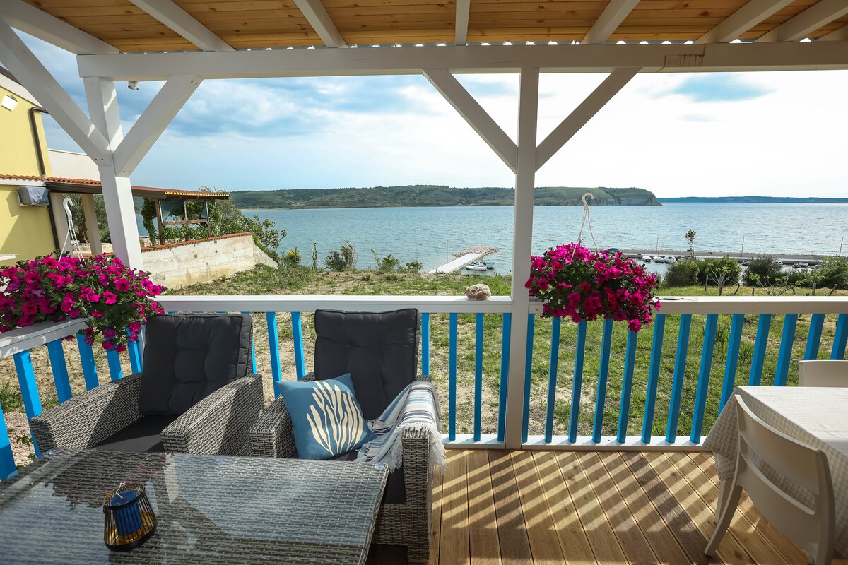 A covered terrace overlooks the sea, featuring a glass-top table surrounded by comfortable wicker seating. Potted flowers in vibrant colors hang from the railing, enhancing the outdoor space. A tranquil view of the water and distant greenery is visible beyond the terrace.