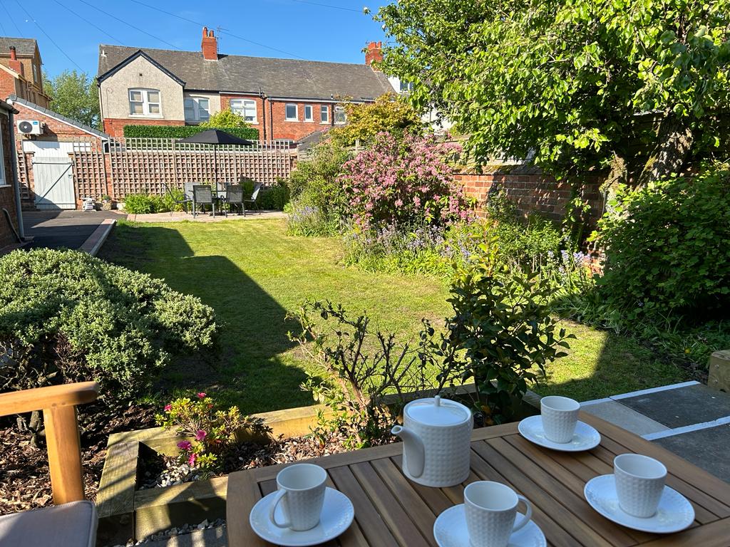 A large back garden is showcased with blooming greenery and flowers. A wooden dining table is set with four white cups. Sunlight casts gentle shadows, highlighting the various plants and a well-maintained lawn, creating a peaceful outdoor space.