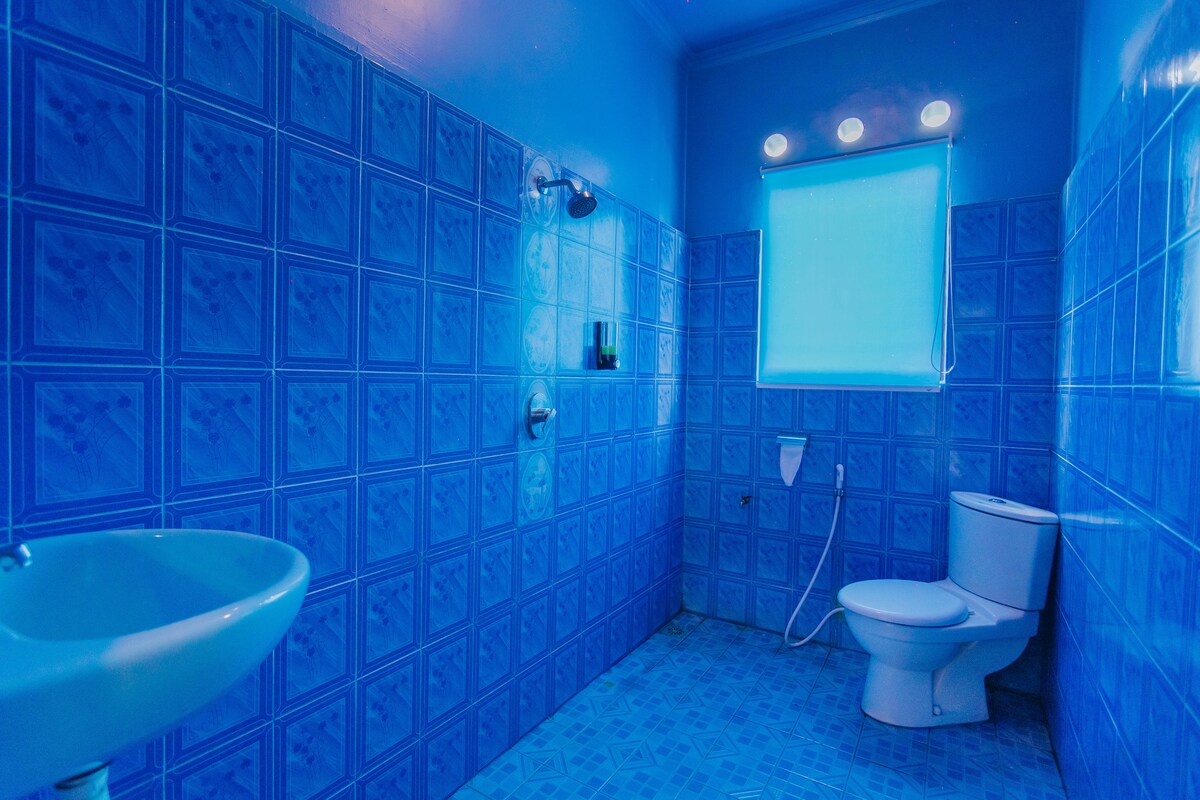 A bright bathroom features blue tiled walls and flooring. A sink with a modern faucet is positioned on one side, while a toilet and shower are placed against the opposite wall. A frosted window allows natural light to enter.