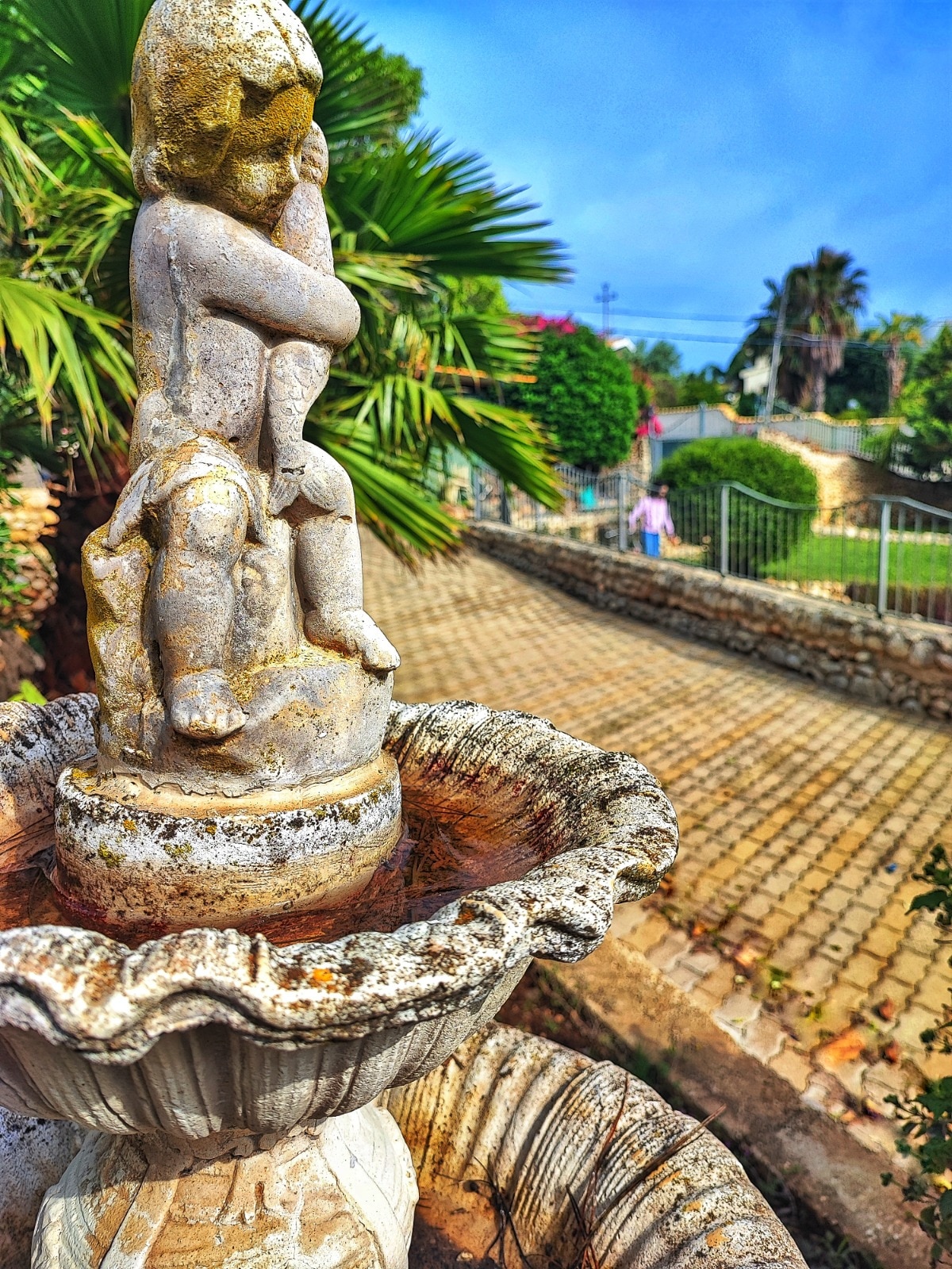 A classic stone fountain features a cherub figure sitting atop a scalloped basin, surrounded by lush greenery. The gently worn surface of the fountain suggests age, while the cobblestone pathway leads into the distance, providing a sense of the garden's spaciousness.