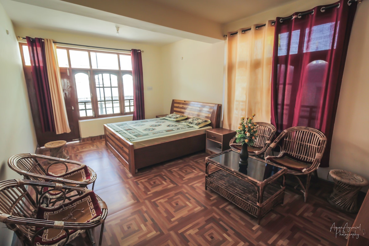 A spacious room features a double bed with a patterned bedspread, surrounded by natural light from three large windows. Two bamboo chairs and a glass-top coffee table are arranged in the seating area, enhancing comfort and accessibility.