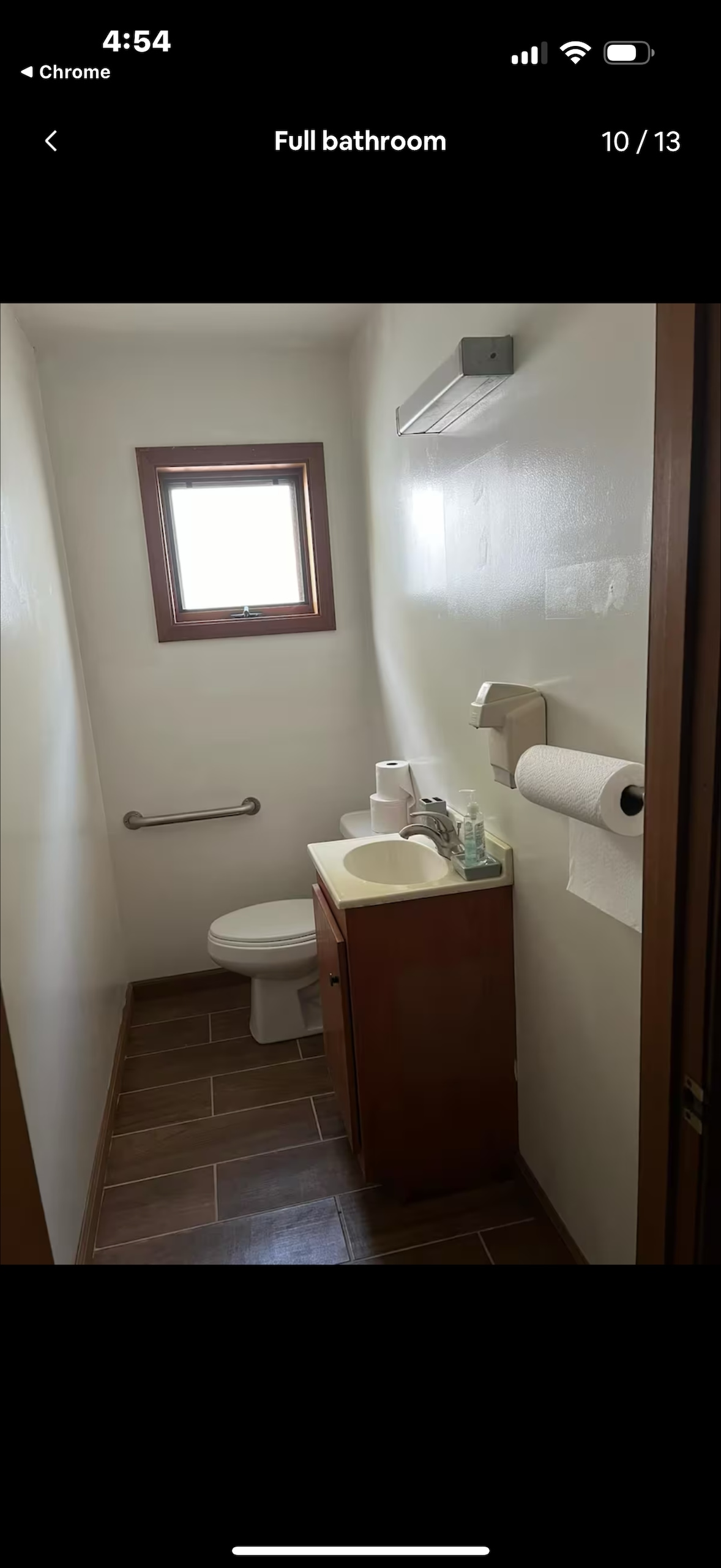 A full bathroom is displayed, featuring a toilet positioned beside a compact vanity with a sink. A window allows natural light to enter the space. Hand towels and toiletries are neatly arranged on the counter, while a grab bar is installed near the toilet for accessibility.