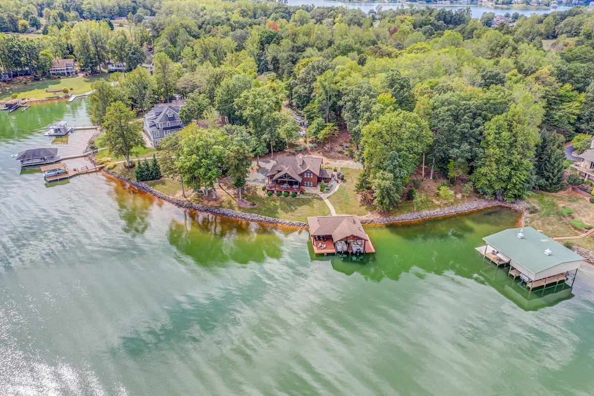 An aerial view showcases the lodge nestled among lush greenery, adjacent to a shimmering lake. The surrounding area features multiple docks, providing easy access to the water. A mix of residential properties and natural landscapes contribute to the scenic setting.
