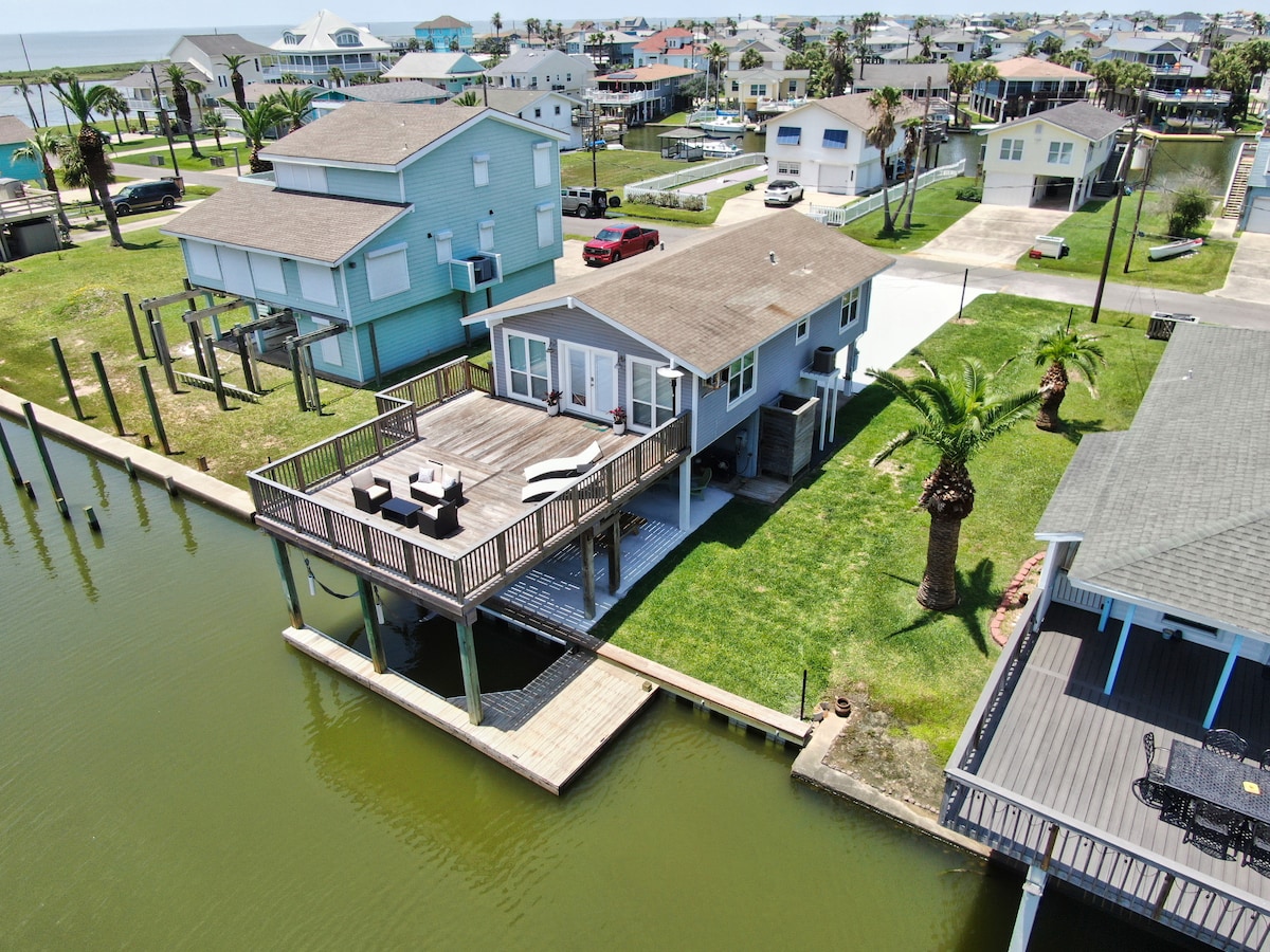 A waterfront home is featured with a spacious deck overlooking the canal. Comfortable outdoor seating is positioned on the deck, offering a relaxing view. Palm trees and grassy areas surround the property, with neighboring houses visible across the water.