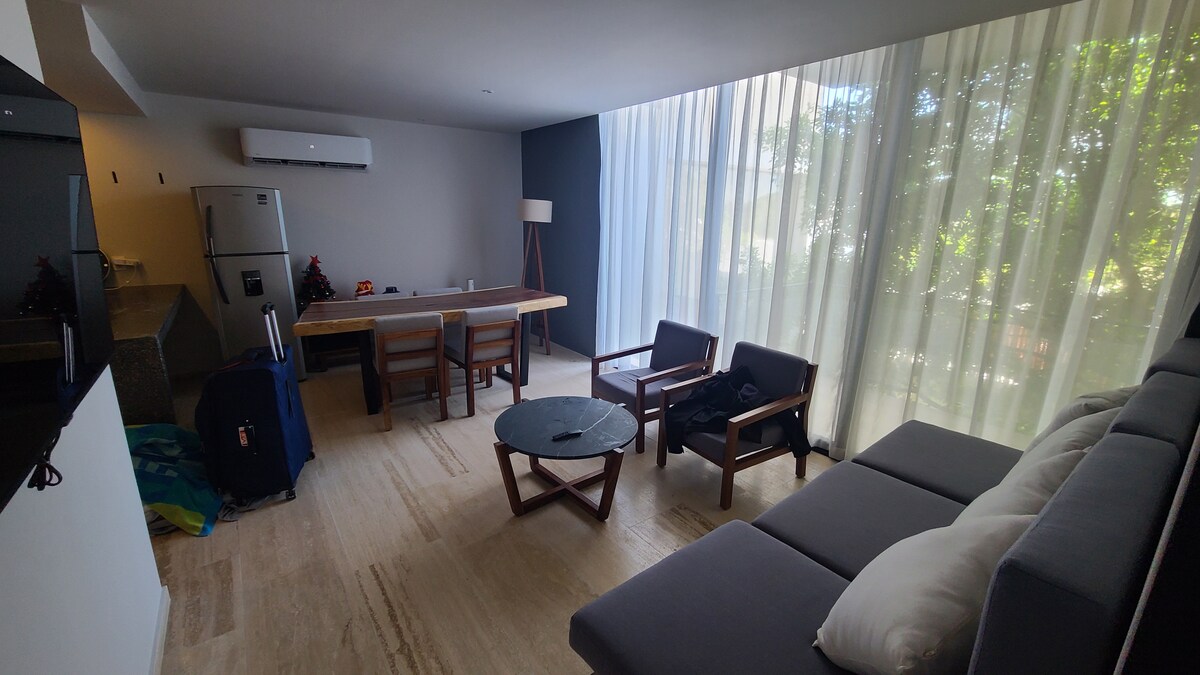 A modern living space features large windows draped with light sheer curtains, allowing natural light to fill the room. A wooden dining table is positioned centrally, accompanied by wooden chairs. A gray sofa is placed facing a circular coffee table, creating a cozy seating area.