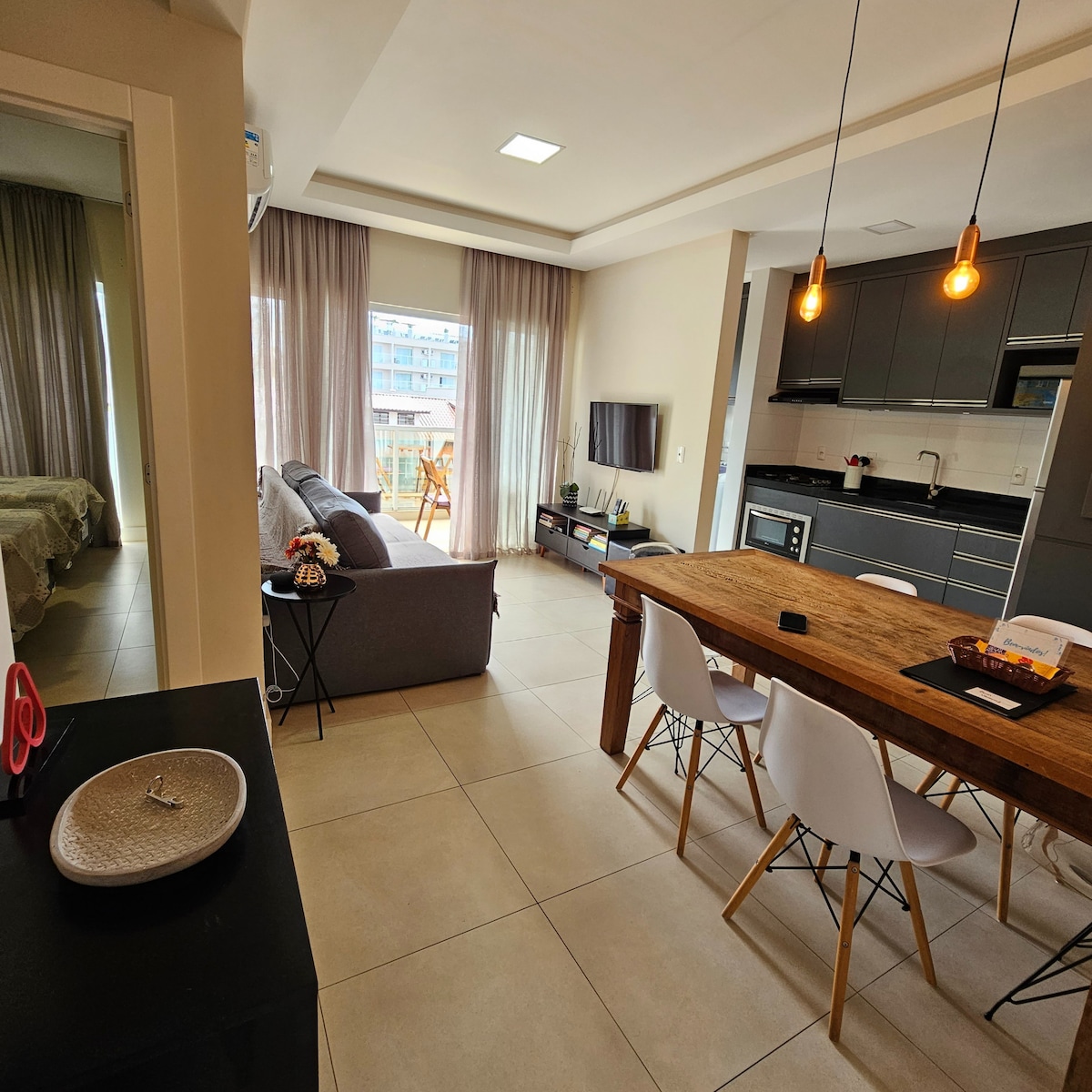 A modern open-concept living and dining area features a wooden dining table surrounded by white chairs. A cozy gray sofa sits opposite a mounted television. Large windows allow natural light to fill the space, complemented by light, flowing curtains.