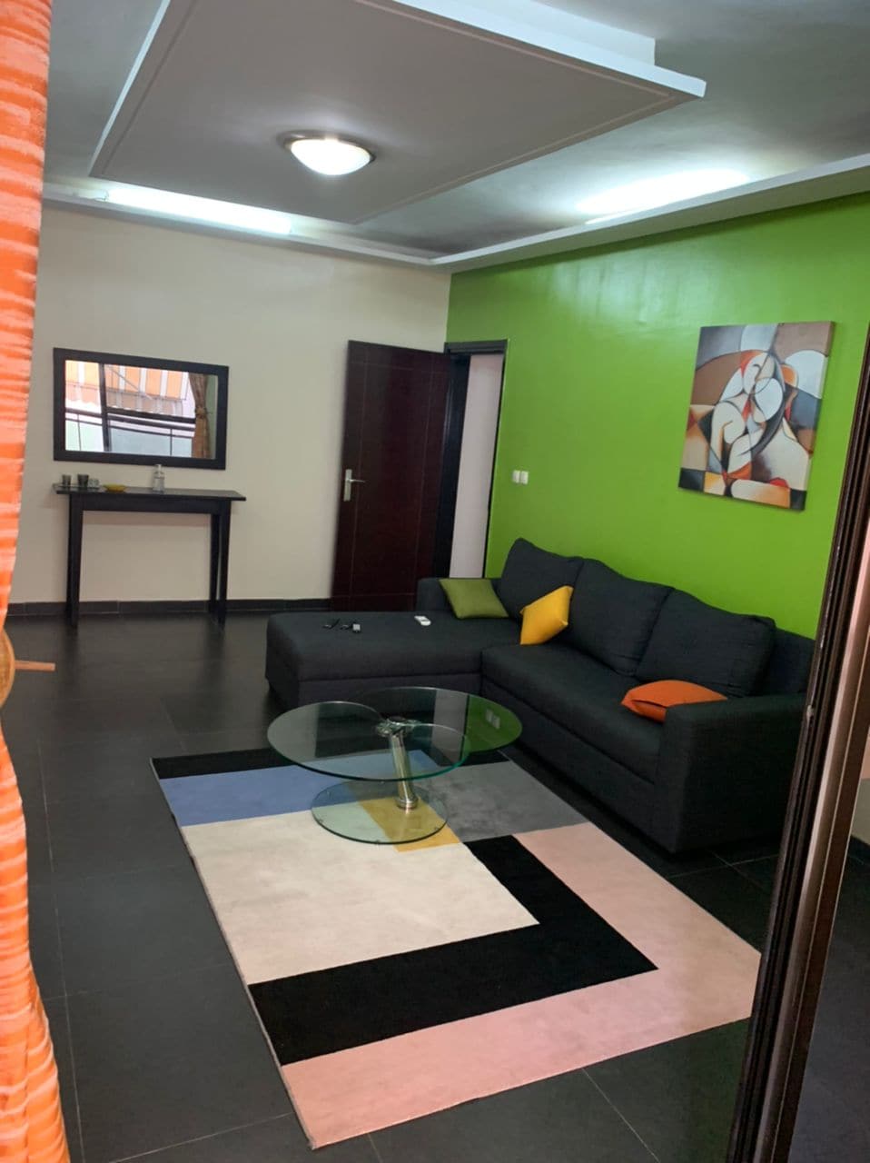 A cozy living area features a black sectional sofa with colorful accent pillows. A circular glass coffee table sits on a patterned rug, while a vibrant green wall adds a lively touch. Natural light is filtered through a nearby window, enhancing the space's brightness.