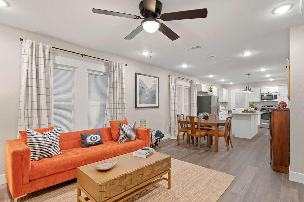 An inviting living space features a vibrant orange sofa paired with decorative pillows. A wooden coffee table is centered on a woven area rug. Natural light fills the room through large windows, complementing the neutral tones of the walls and curtains. A dining area is visible in the background.