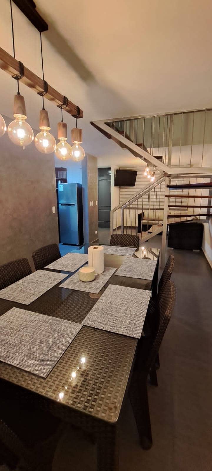 A dining area is highlighted by a large glass table surrounded by dark wicker chairs. The space is illuminated by pendant lights, and a stylish staircase leads to an upper level. A kitchen is visible in the background.