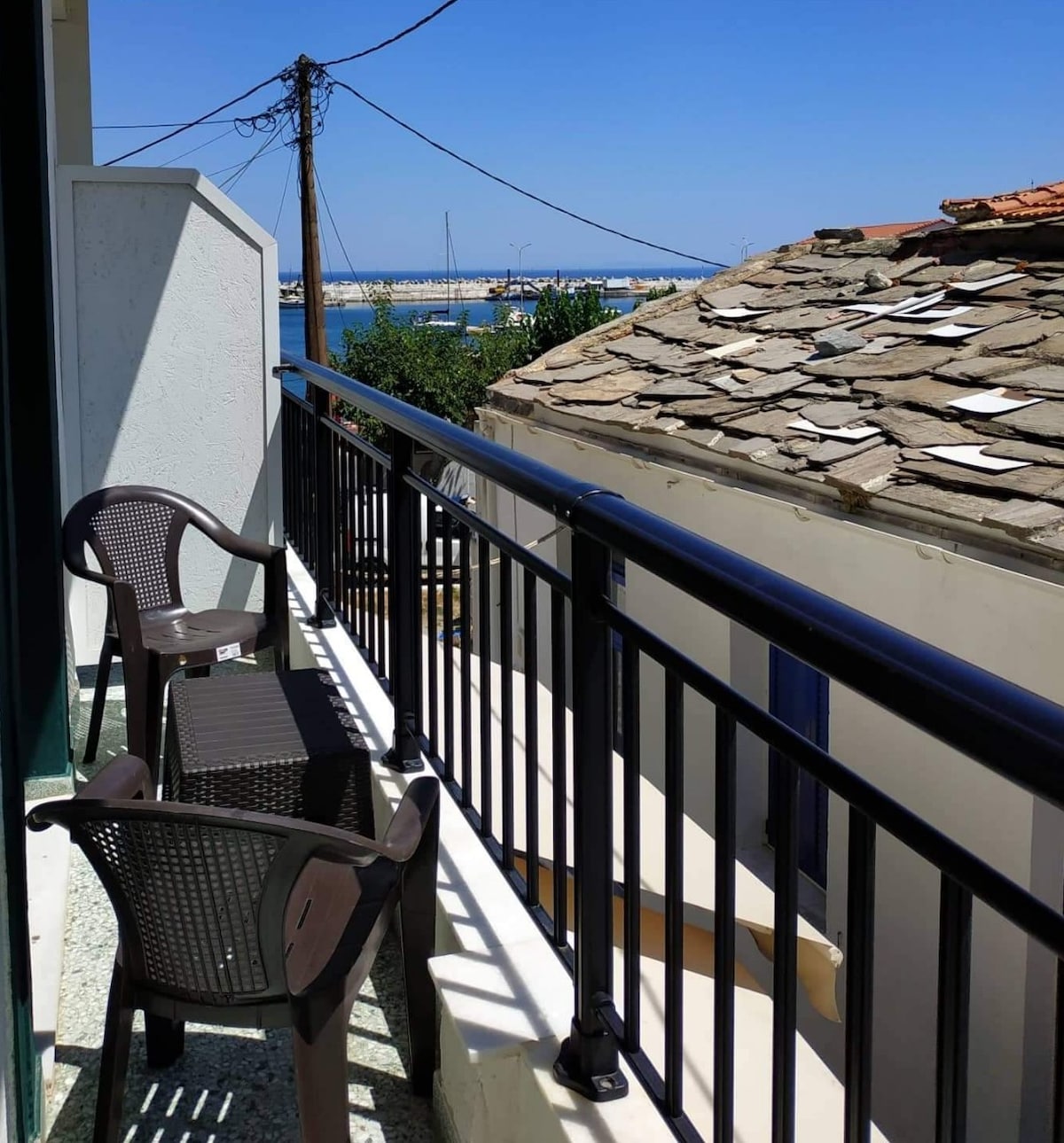 A balcony overlooking the sea features two black plastic chairs and a small table. The view includes distant boats and a clear blue sky. Nearby, a sloped roof showcases rustic tiles, adding texture to the scene.