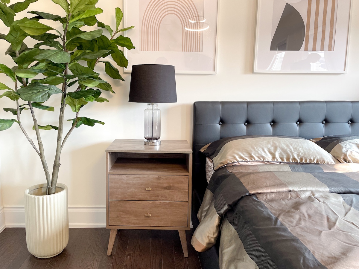 The sleeping area features a comfortable double-size bed with textured bedding and a stylish headboard. A bedside table with two drawers holds a sleek lamp, while a decorative indoor plant adds a touch of greenery. Wall art enhances the modern aesthetic.