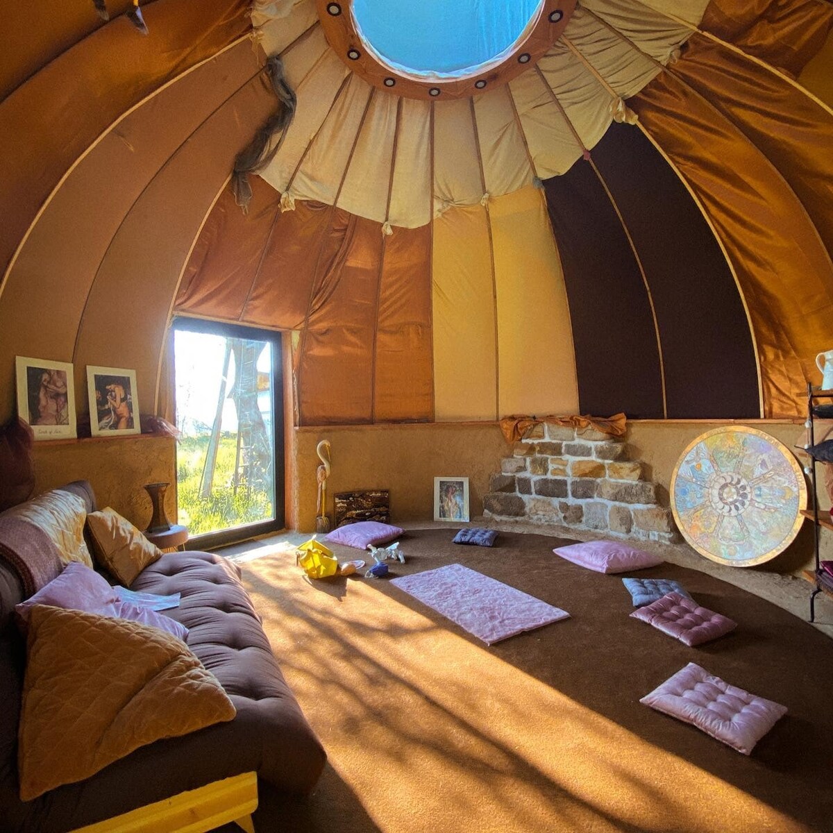 The interior of a dome-shaped structure features warm-toned fabric walls and a circular skylight. A low, comfortable seating area is surrounded by cushions on a soft floor. A stone fireplace is visible, along with framed artwork and a decorative circular piece leaning against the wall.