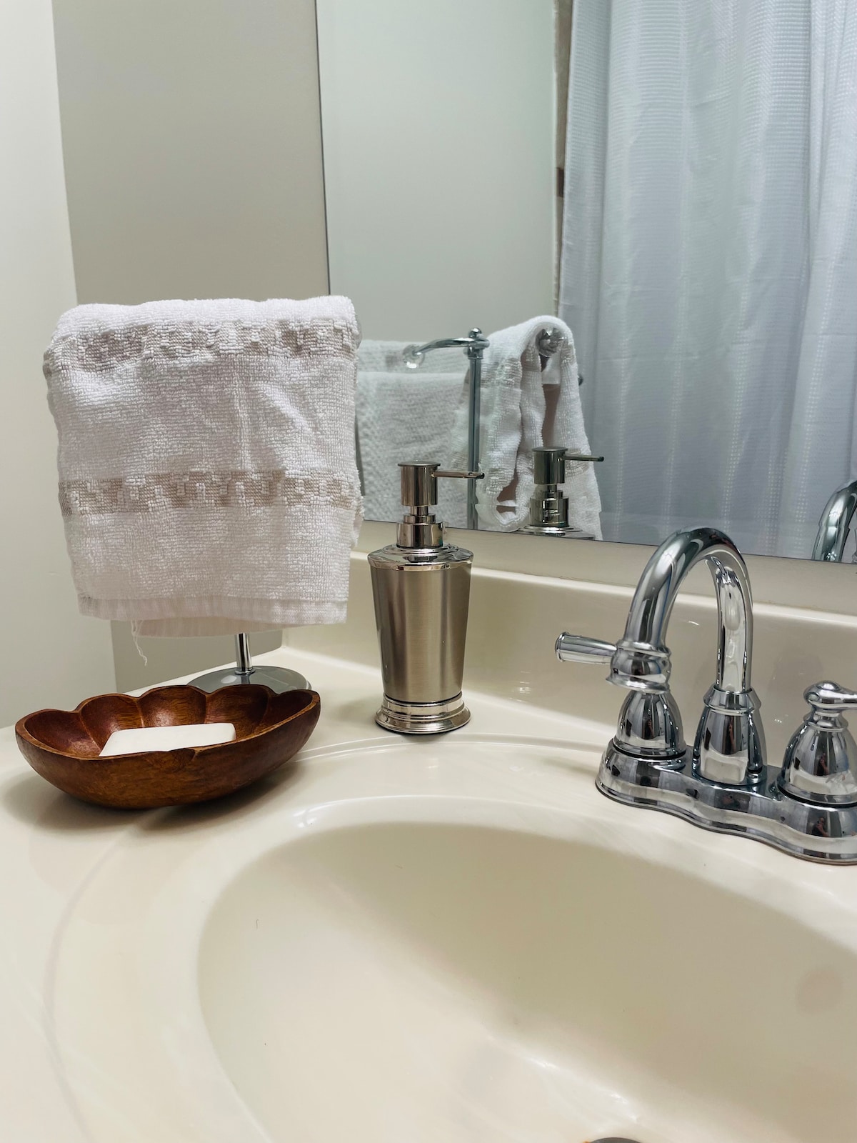 A bathroom sink area is shown, featuring a smooth countertop with a bowl containing a bar of soap. A silver soap dispenser is positioned nearby. Clean white towels hang on a rack, and a mirror reflects the space, with a light-colored shower curtain visible in the background.