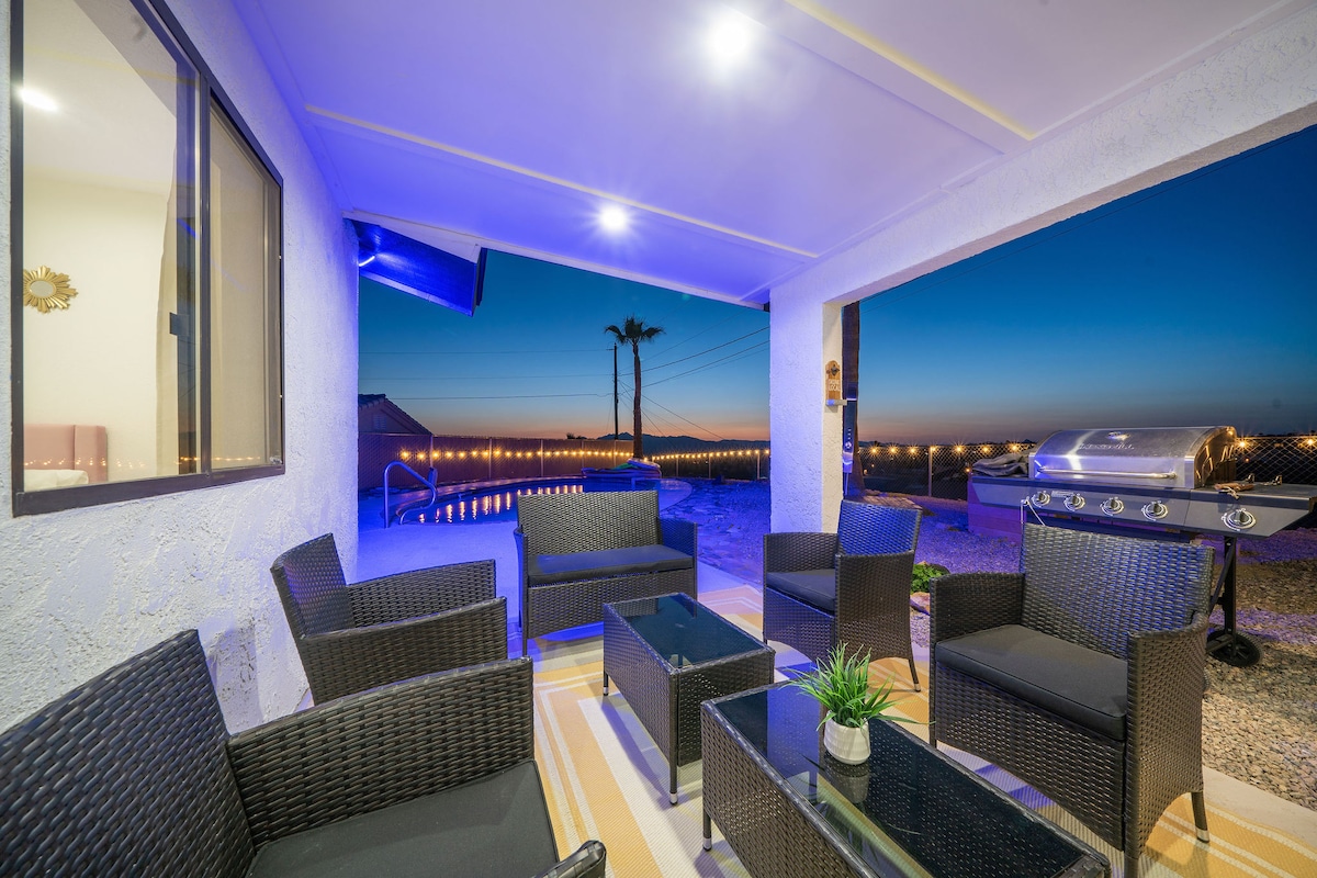 An outdoor seating area features comfortable dark wicker chairs and a glass-top table, set against a backdrop of a twilight sky. A gas grill stands nearby, and the inviting pool glimmers in the distance, creating a serene atmosphere for relaxation.