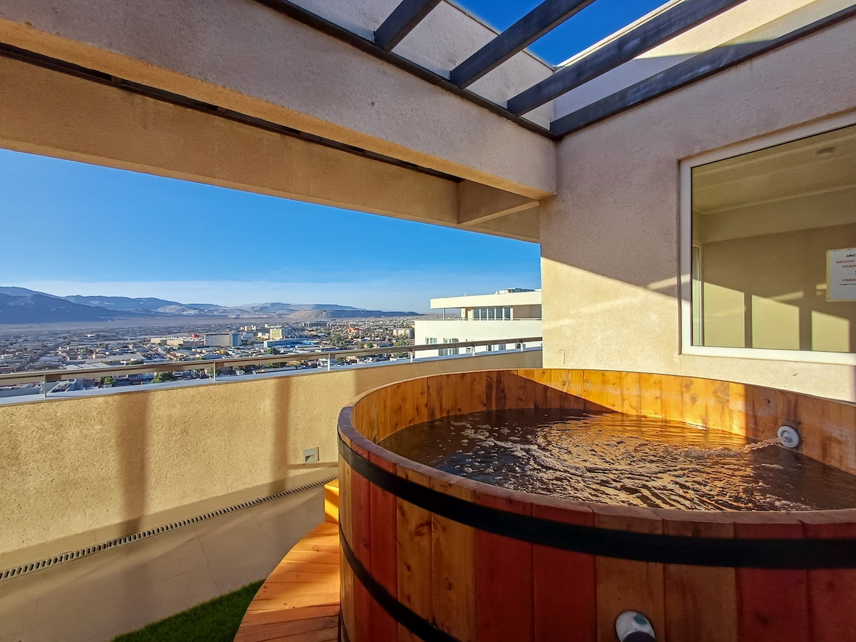 A wooden hot tub is positioned on a private balcony, offering unobstructed views of the surrounding landscape. Sunlight reflects off the water's surface, and the space is partly covered by clear panels, allowing for an open yet sheltered experience.