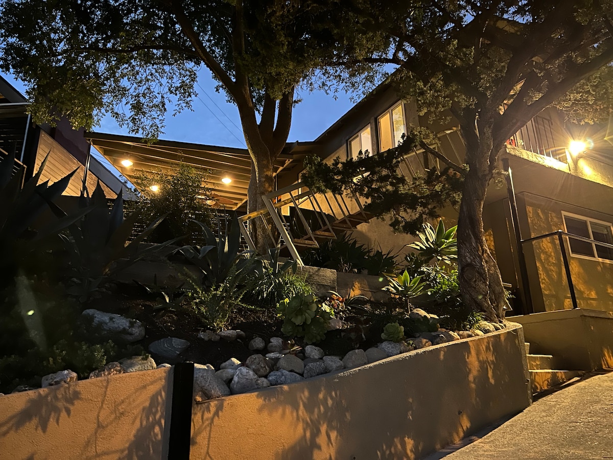 The hillside guest suite is surrounded by a well-maintained garden featuring varied foliage and decorative stones. Soft lighting highlights the studio's entrance, providing a welcoming atmosphere at dusk. A textured pathway leads up to the suite, bordered by lush plants and trees.