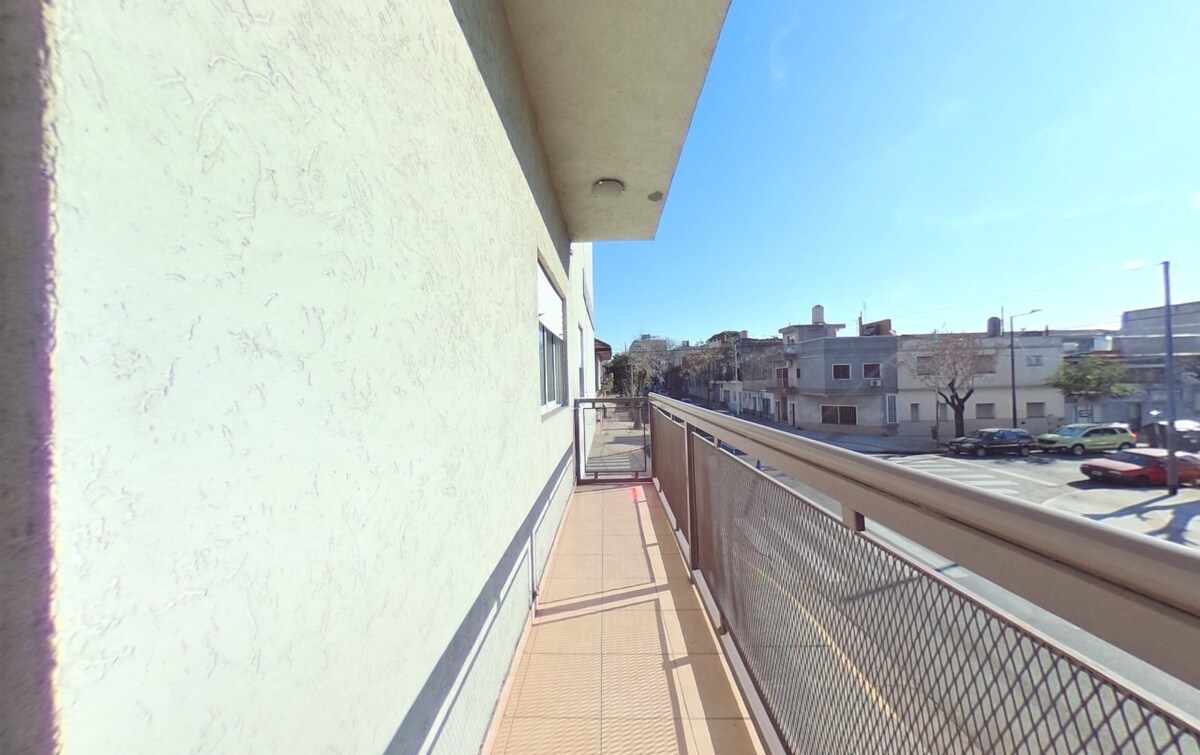 A spacious balcony is visible, extending along the side of the building. The floor is tiled, and a railing is present. A clear view of the street below includes buildings and parked cars, with sunlight illuminating the area.