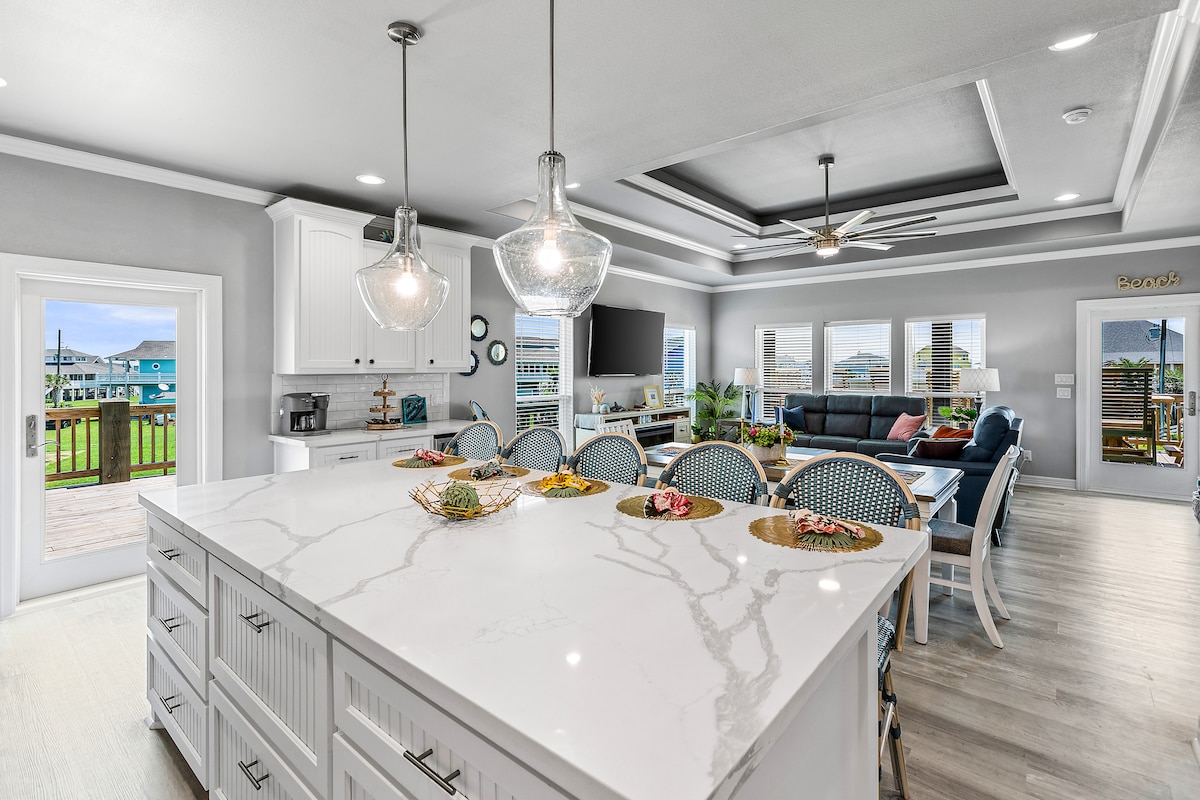 The kitchen features a large, white island with seating, surrounded by modern appliances and cabinetry. Natural light fills the space through multiple windows, showcasing views of the outdoor area. A dining area with a comfortable seating arrangement is visible in the background.