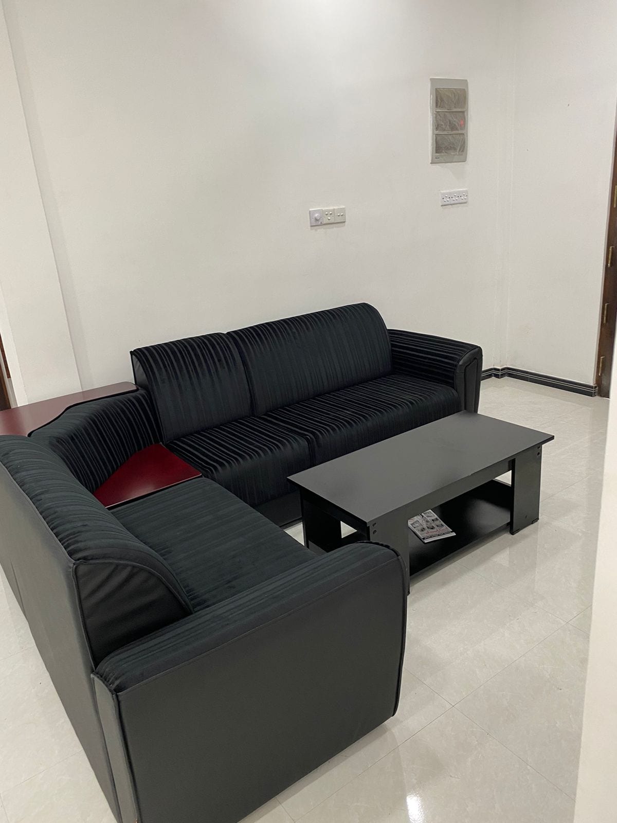 A comfortable seating area is featured with a black-striped sofa set arranged in an L-shape. A dark coffee table is placed in the center, alongside a remote control. The walls are painted white, contributing to a clean and minimalistic appearance.