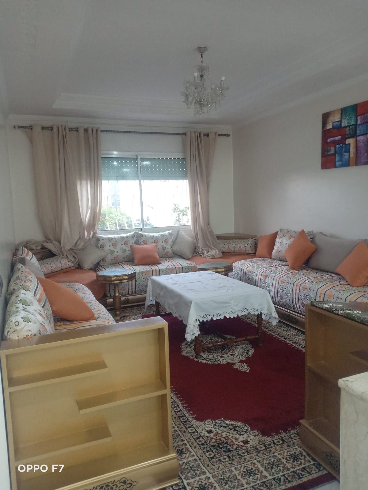 A traditional Moroccan living area features multiple patterned sofas accented with orange cushions, arranged around a low table. A colorful wall art piece adds character, while natural light filters through sheer curtains covering the window, enhancing the serene atmosphere.