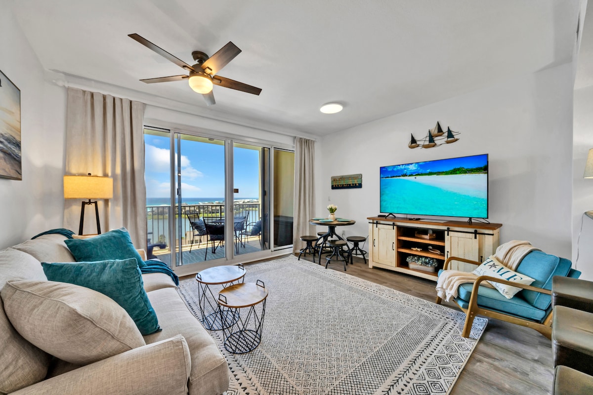 The living area features a comfortable sofa accompanied by teal accent pillows, with a light rug beneath. A large television is mounted on a wooden stand, while a ceiling fan adds to the airiness. Natural light pours in from the balcony doors, enhancing the welcoming atmosphere.