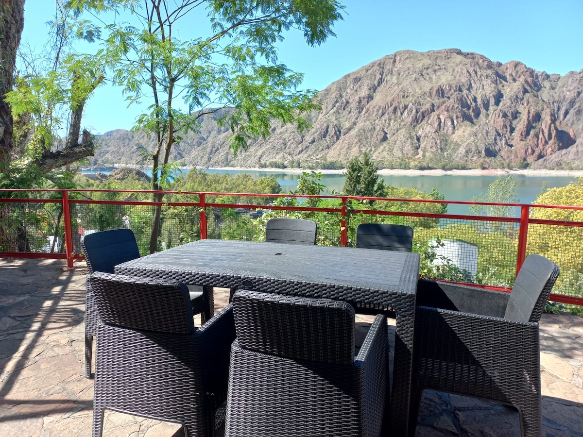 A dining area with a large, dark wicker table is surrounded by six matching chairs. The space is framed by trees, offering views of the tranquil lake and rocky hills in the background under a clear blue sky.