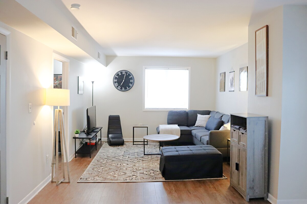 A light-filled living area features a comfortable sectional sofa and a round coffee table. A large wall clock is displayed prominently above the TV stand, enhancing the modern decor. Artwork adorns the walls, and a patterned rug adds warmth to the space.