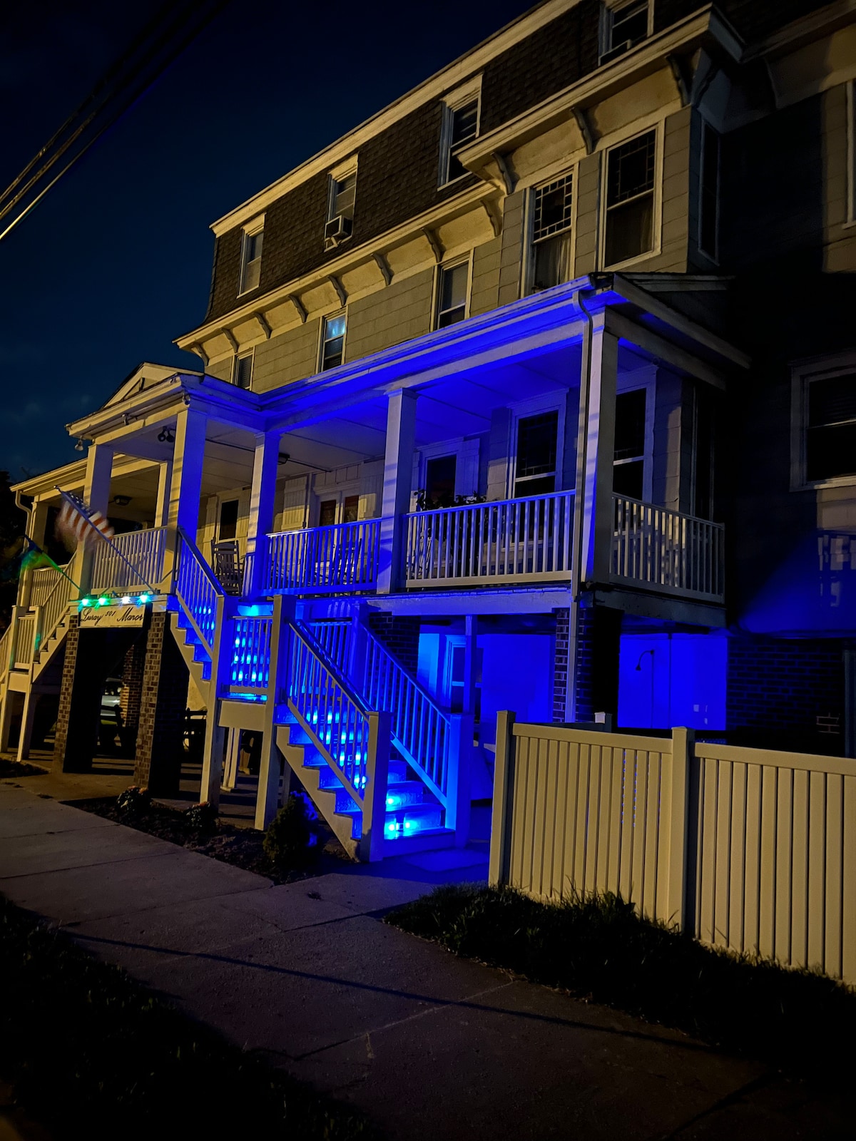 A multi-story building is illuminated by blue lights, highlighting its porches and steps. The structure features white railings and a clean exterior. A white fence surrounds the property, enhancing its visibility at night. The street is adjacent to the building.