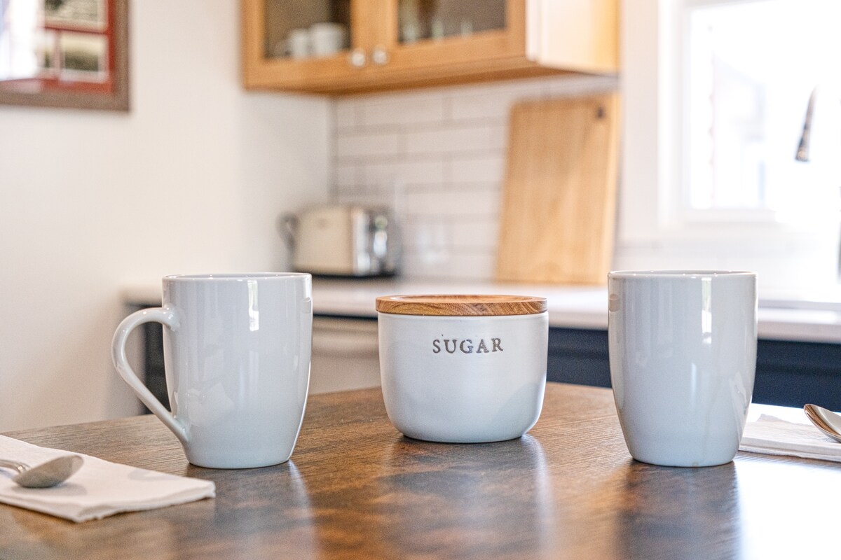 A wooden table displays two matching white mugs and a sugar bowl with a wooden lid. The background features a well-lit kitchen with wood cabinetry and visible kitchen appliances. Bright natural light enters through a nearby window.