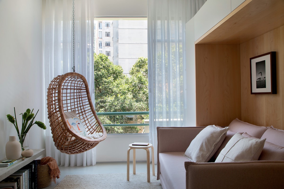 A cozy seating area features a modern sofa with two decorative pillows, complemented by a light-colored stool. A hanging rattan chair adds an inviting touch, while sheer curtains allow natural light to fill the space. Greenery is visible through large windows, enhancing a serene ambiance.